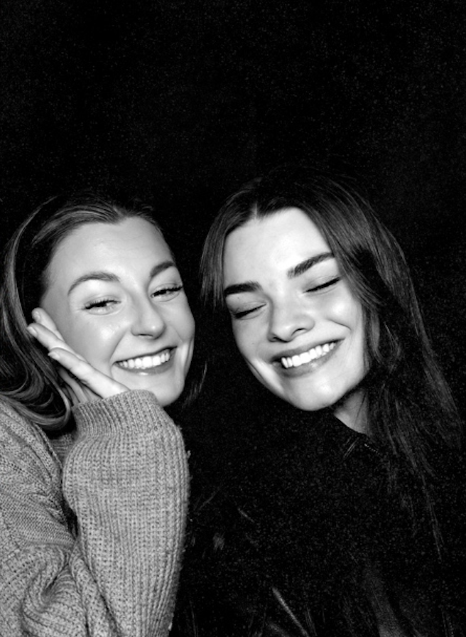 Two women smiling with eyes closed, posing closely together in a black and white photo.