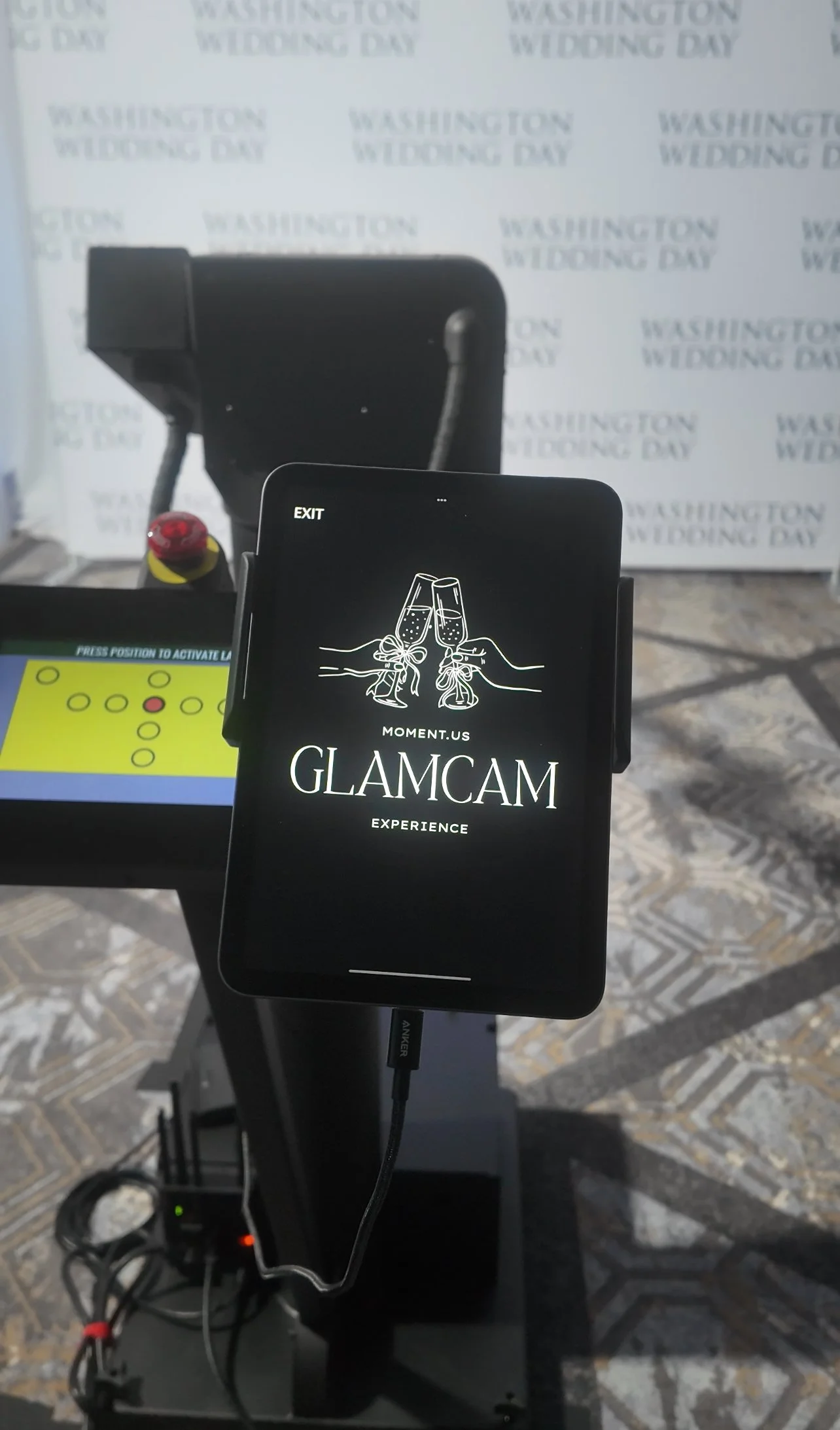 A smartphone on a stand displaying the GLAMCAM experience logo with two champagne glasses clinking, set against a background with washing instructions that read WASHINGTON WEDDING DAY.