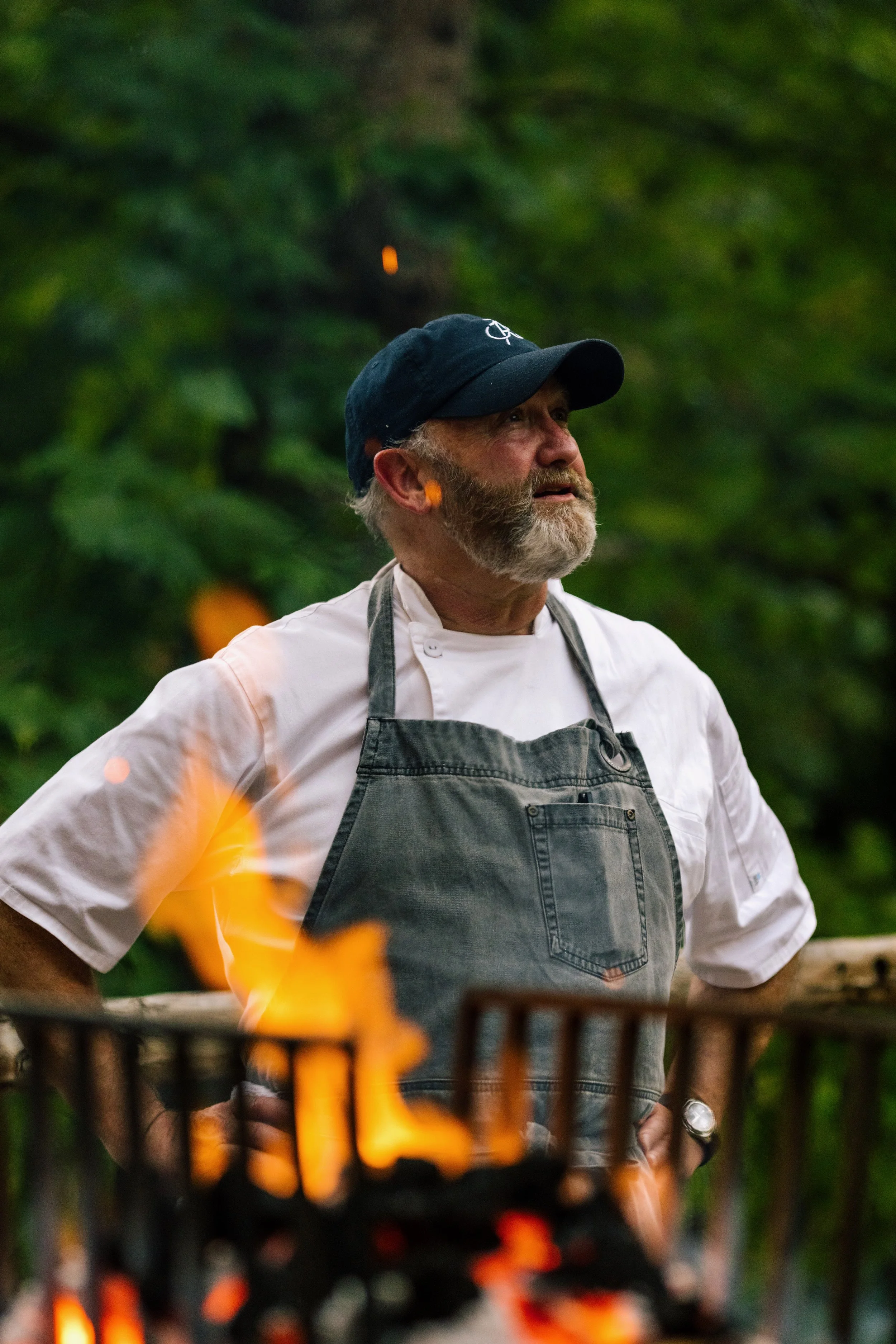 Chef wearing a hat and apron standing near a grill with visible flames, surrounded by greenery.