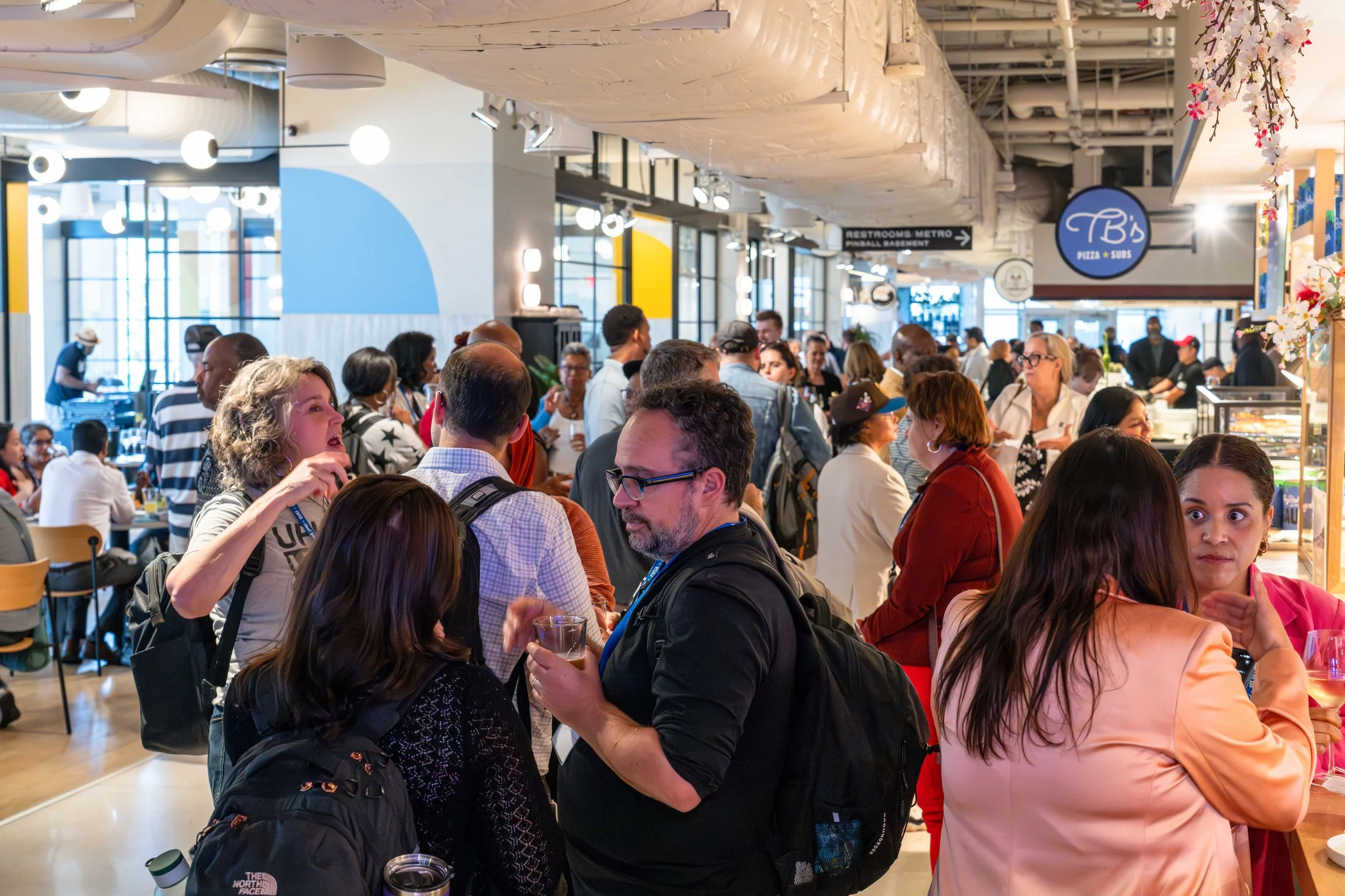Crowded indoor food court or market with many people, some holding drinks, and food stalls in the background, with signs for pizza and subs and directions to restrooms and metro.