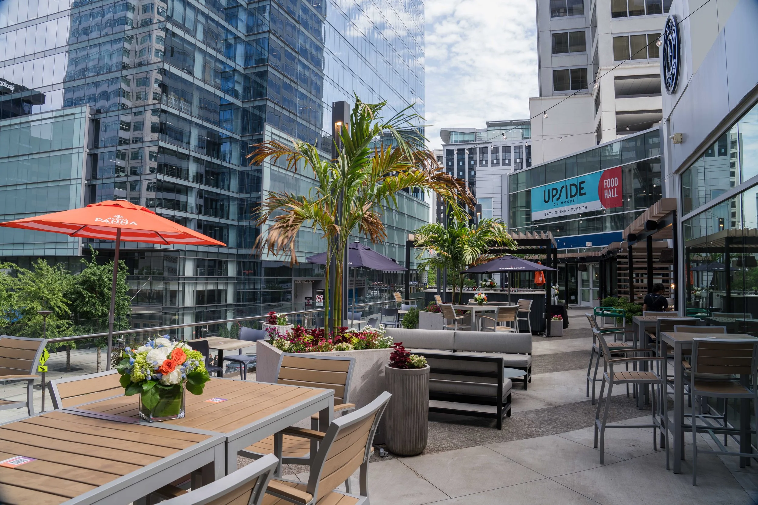 Outdoor urban patio with tables, chairs, and umbrellas, surrounded by modern glass high-rise buildings, with decorative plants and flowers, and a sign for Up & Down restaurant.