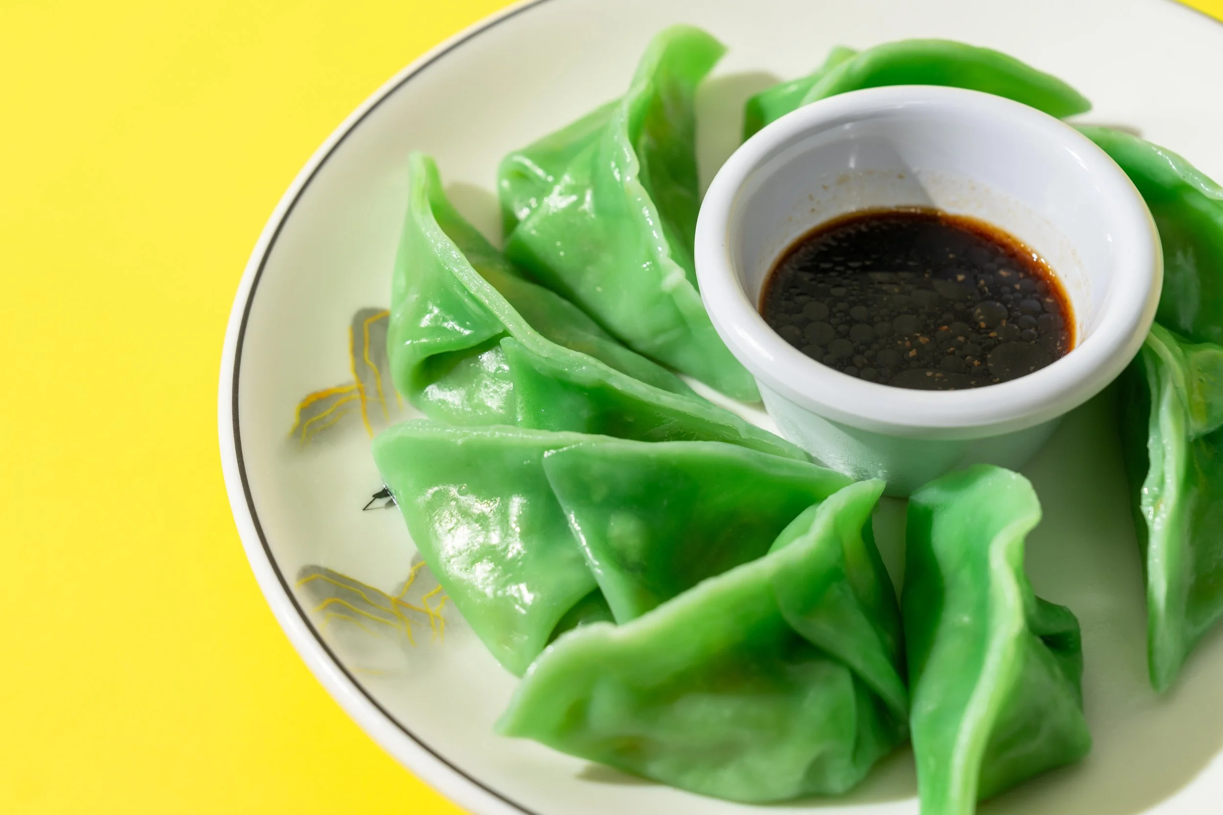 Green dumplings arranged on a white plate with yellow accents, served with soy sauce in a small white bowl, against a yellow background.