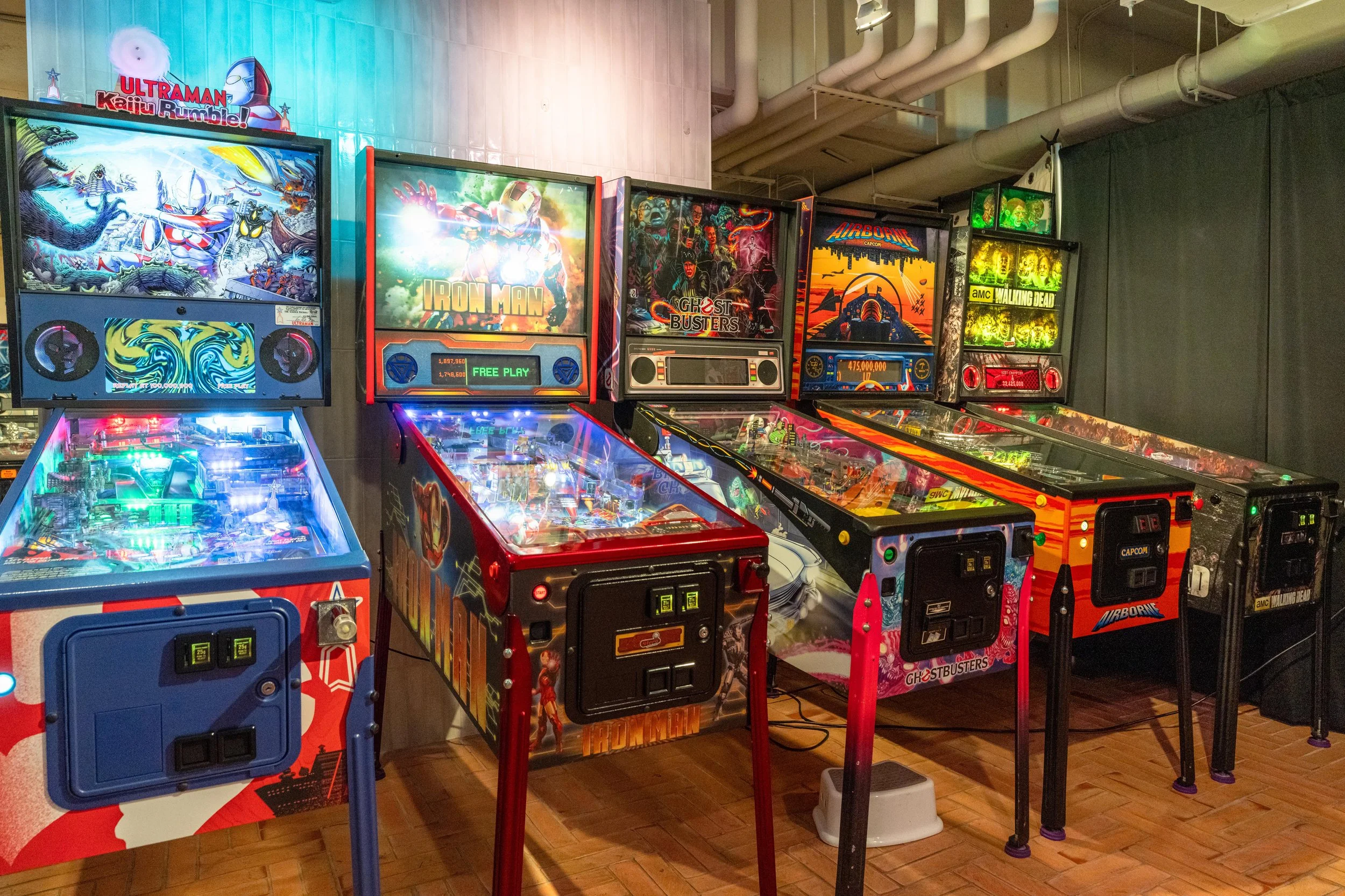 Multiple pinball machines lined up in a game arcade, featuring themes like Ultraman, Iron Man, Ghostbusters, Airborne, and Walking Dead.