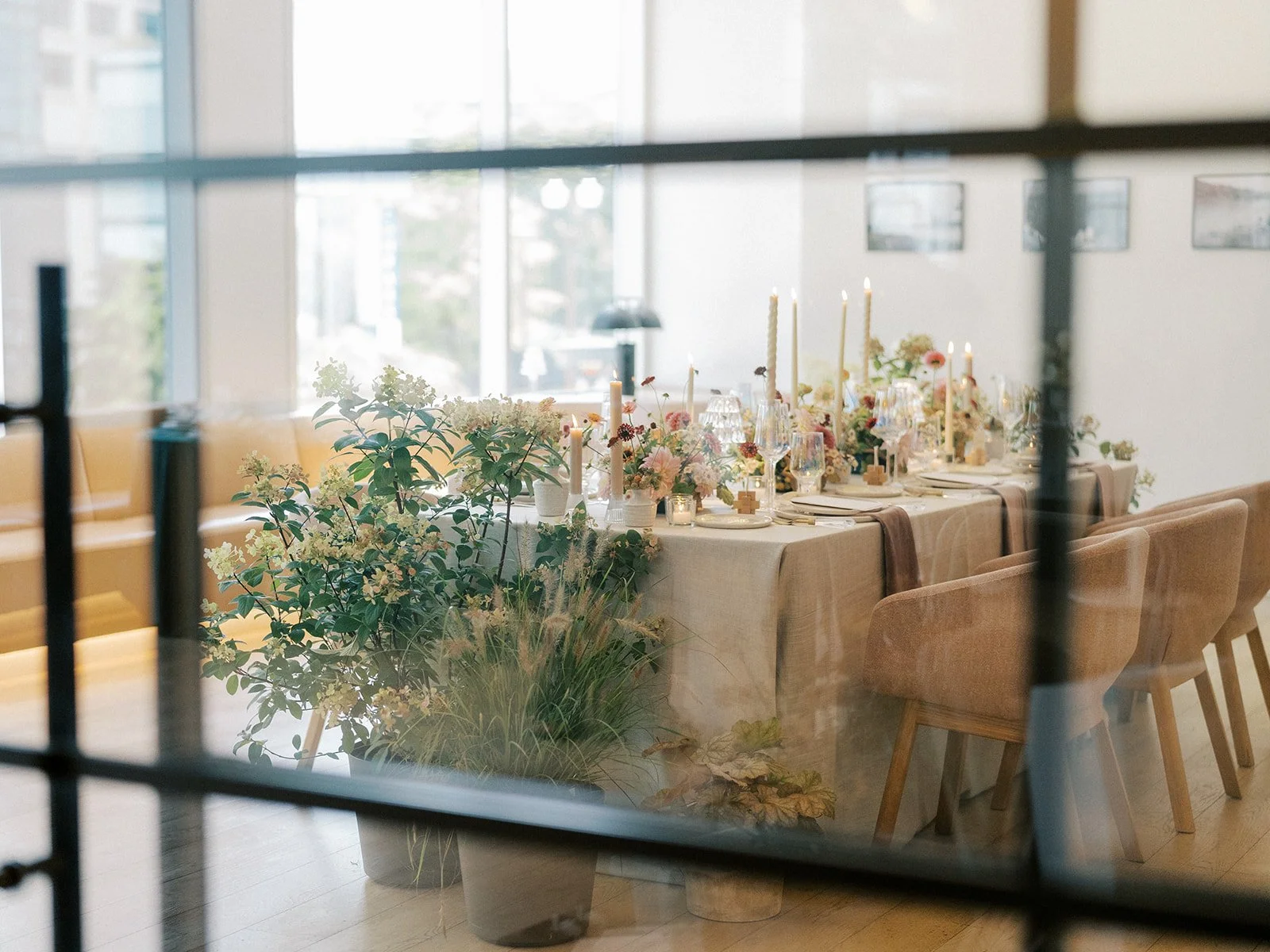 Elegant dining table decorated with flowers, candles, and table settings, seen through a window with black framing.