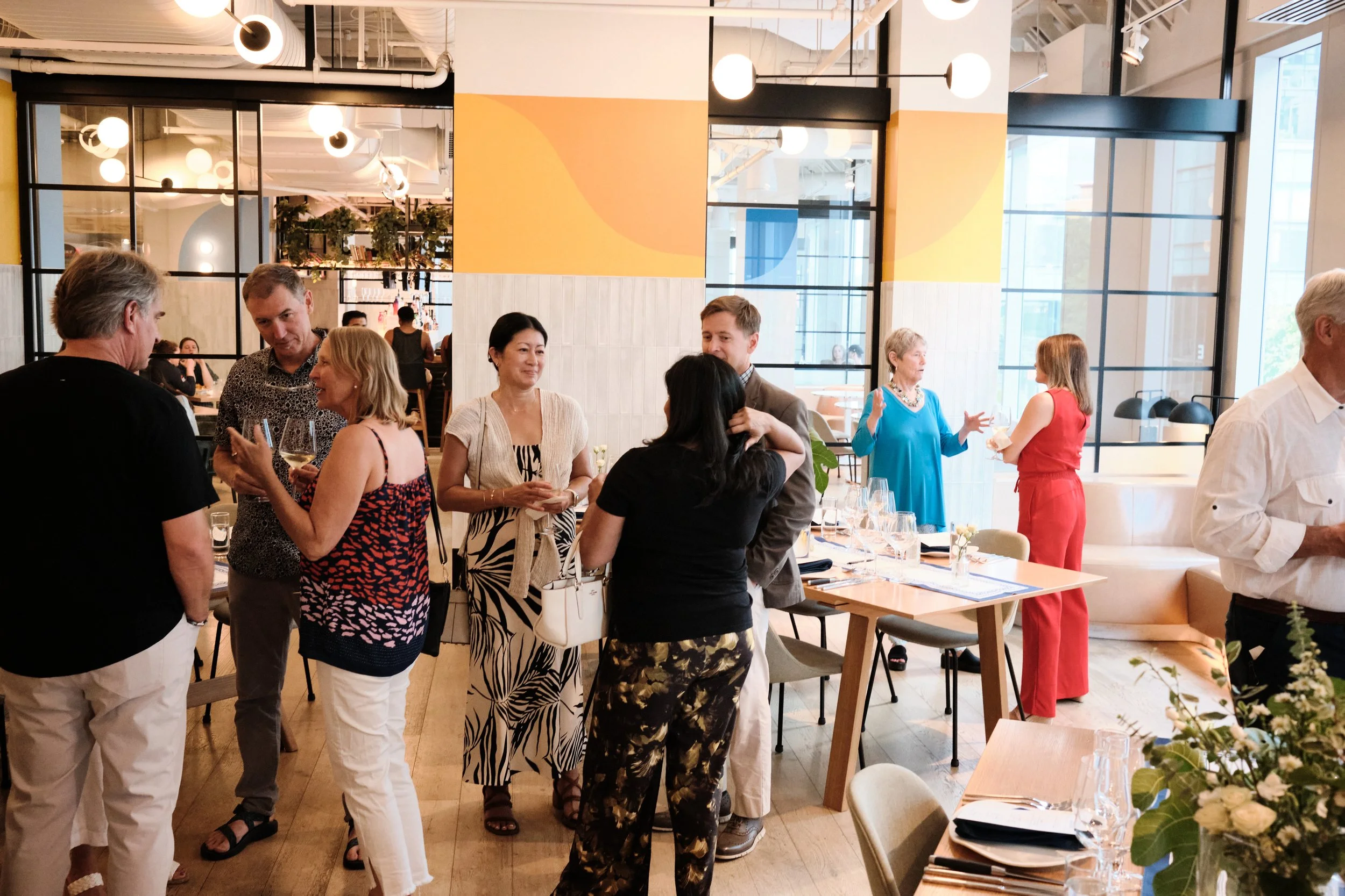 People socializing and talking at a gathering in a brightly lit modern restaurant with large windows and colorful wall art.