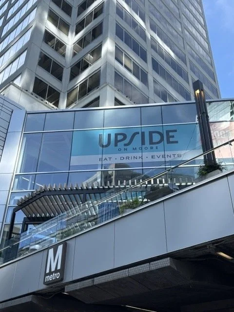 Skyscraper with a glass storefront sign for 'UPSIDE ON MOORE' that offers eat, drink, events, with a metro station entrance labeled 'M' underneath.