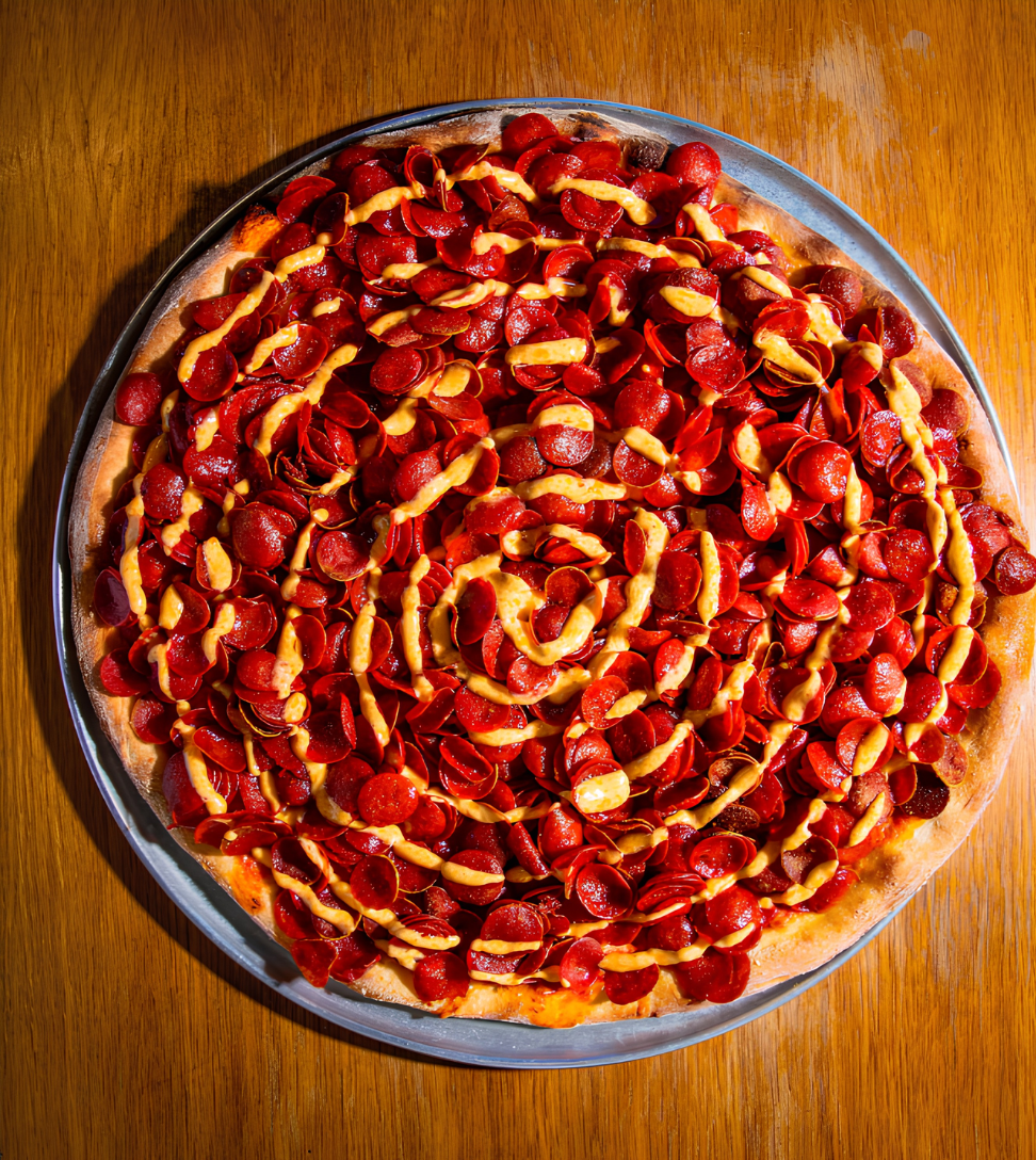 A pizza topped with numerous slices of pepperoni and a drizzle of mustard, placed on a metal tray on a wooden table.