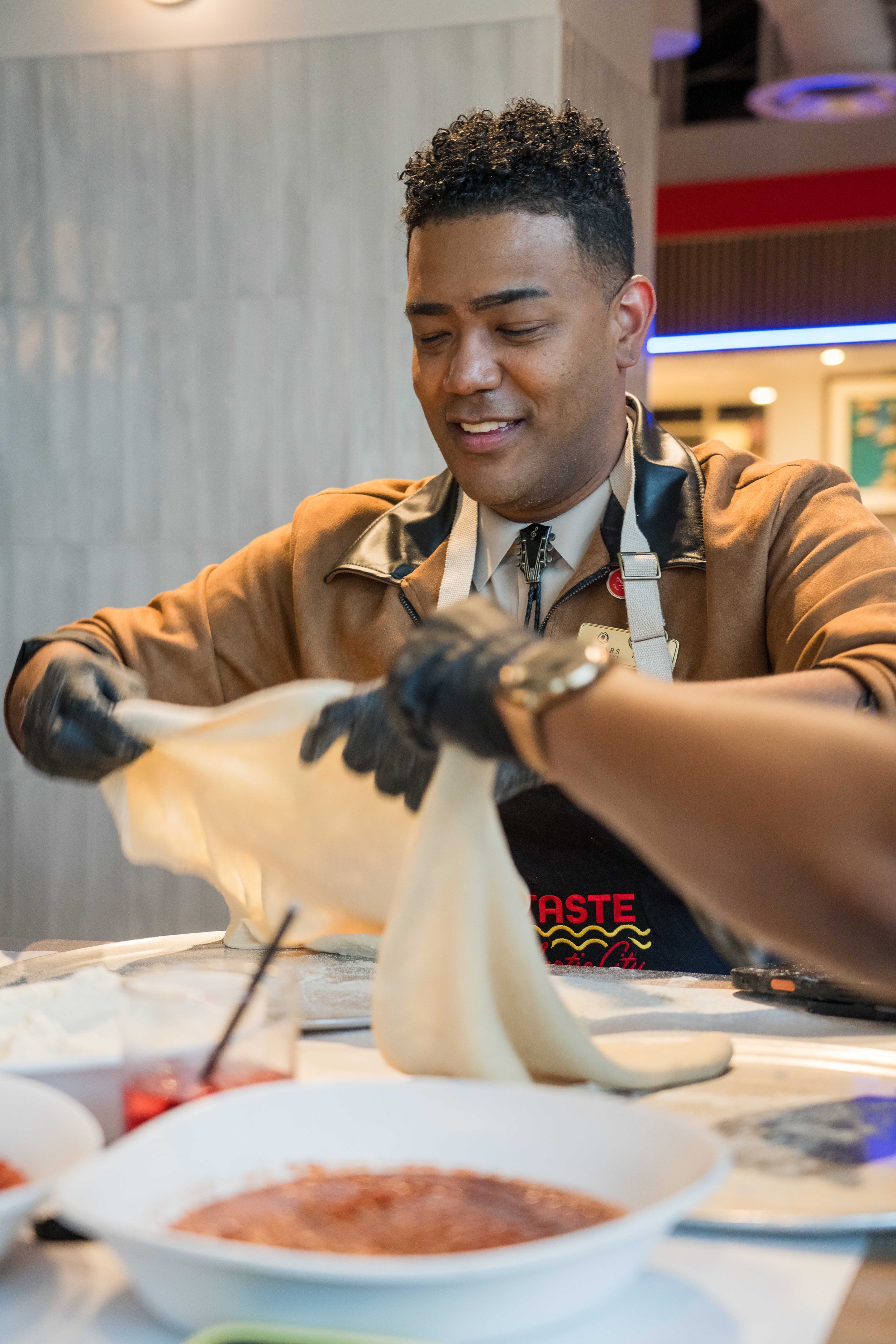 A man with curly hair is smiling and preparing food in a restaurant setting, wearing a brown jacket and black apron with red text.