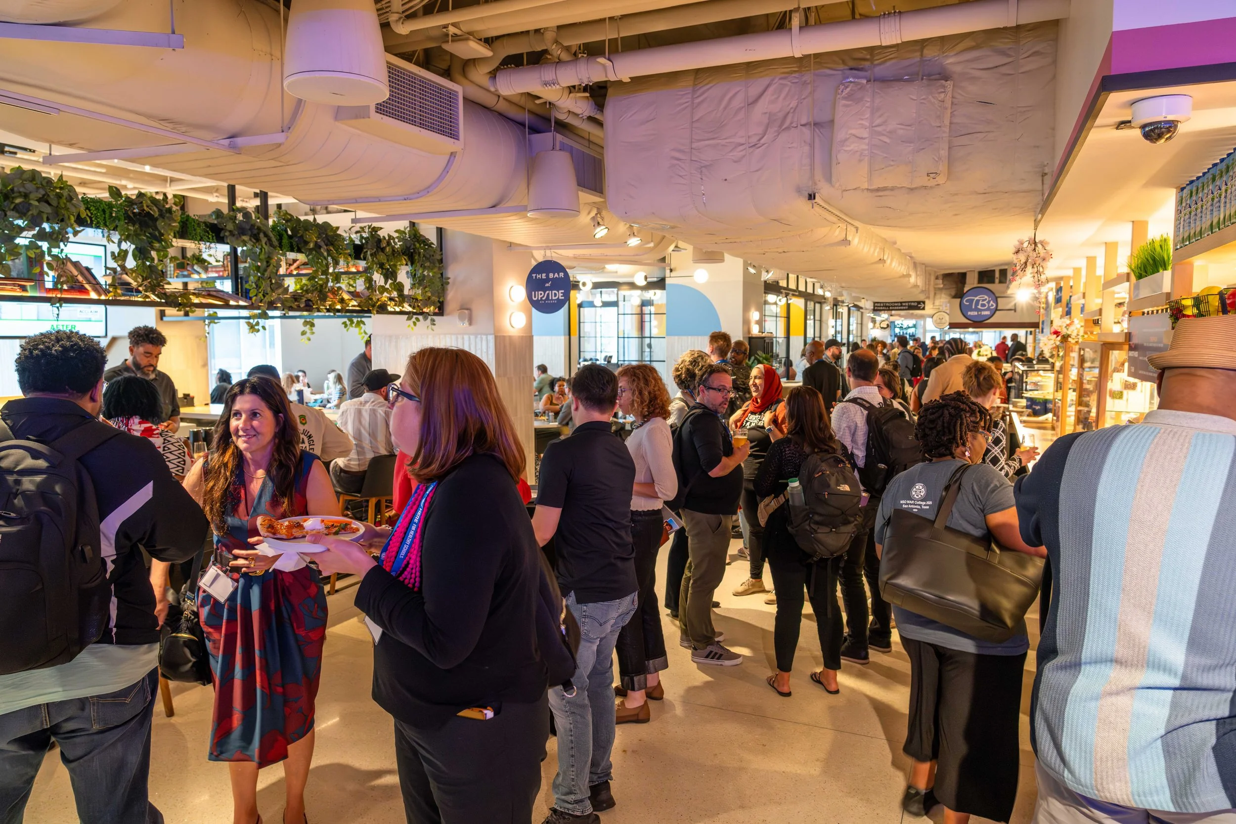 Crowd of people inside a busy food hall or market, some waiting and some eating, with various counters and signs in the background such as 'The Bar at UpRise' and others, modern ceiling with exposed ductwork, plants, and bright lighting.