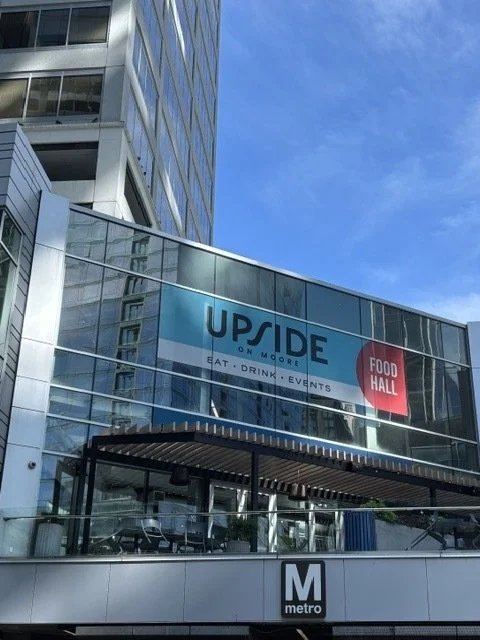 Exterior view of a commercial building with glass windows, a sign for UP SIDE and a food hall, and a metro station entrance in the foreground.