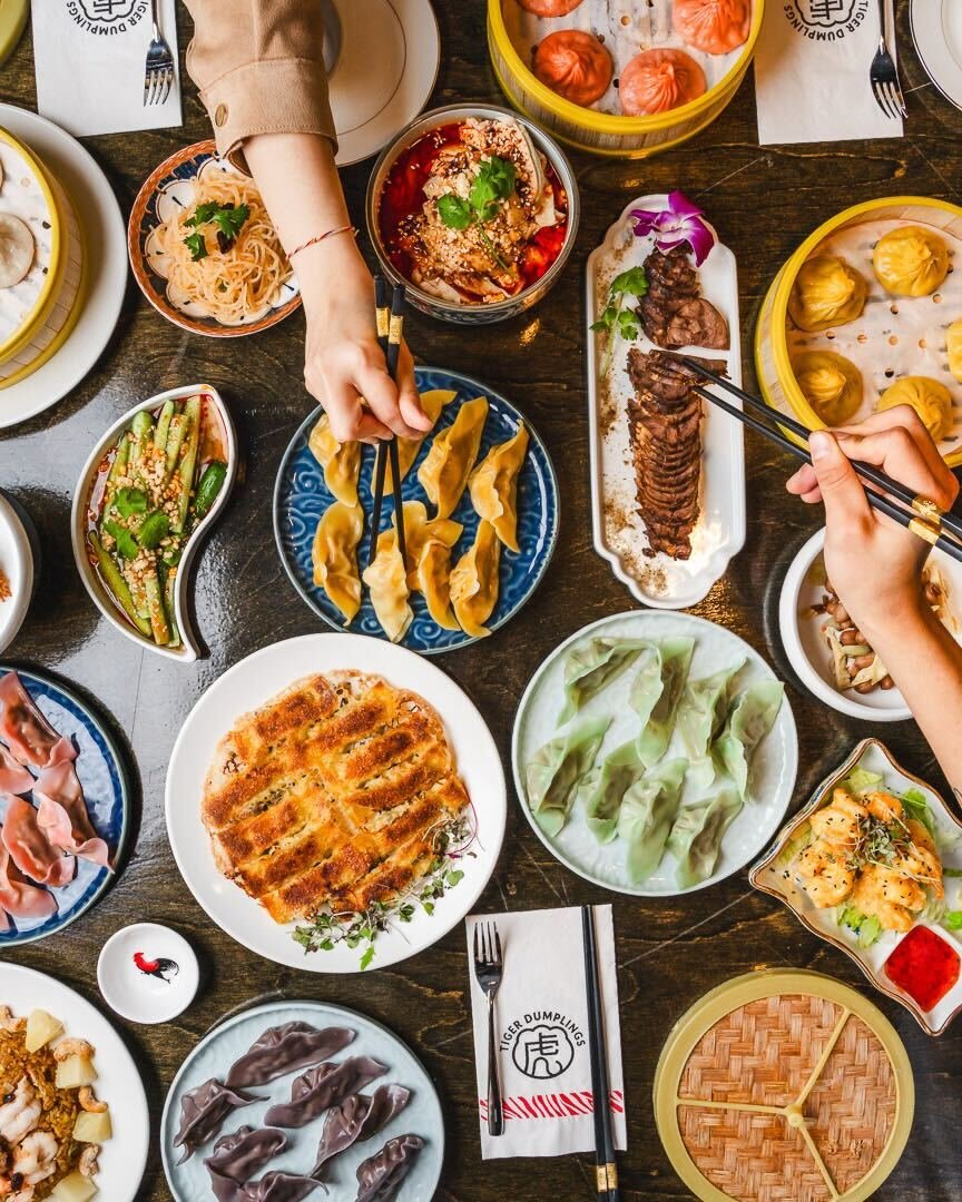 A top-down view of a table filled with various Asian dishes, including dumplings, spring rolls, noodles, and vegetables, with people's hands using chopsticks to pick up food.