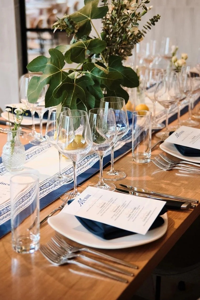 A elegantly set dining table with clear wine glasses, water glasses, plates with black napkins, cutlery, and menus. Decorated with a large green leafy plant and smaller flowers in vases, with lemons as centerpieces.