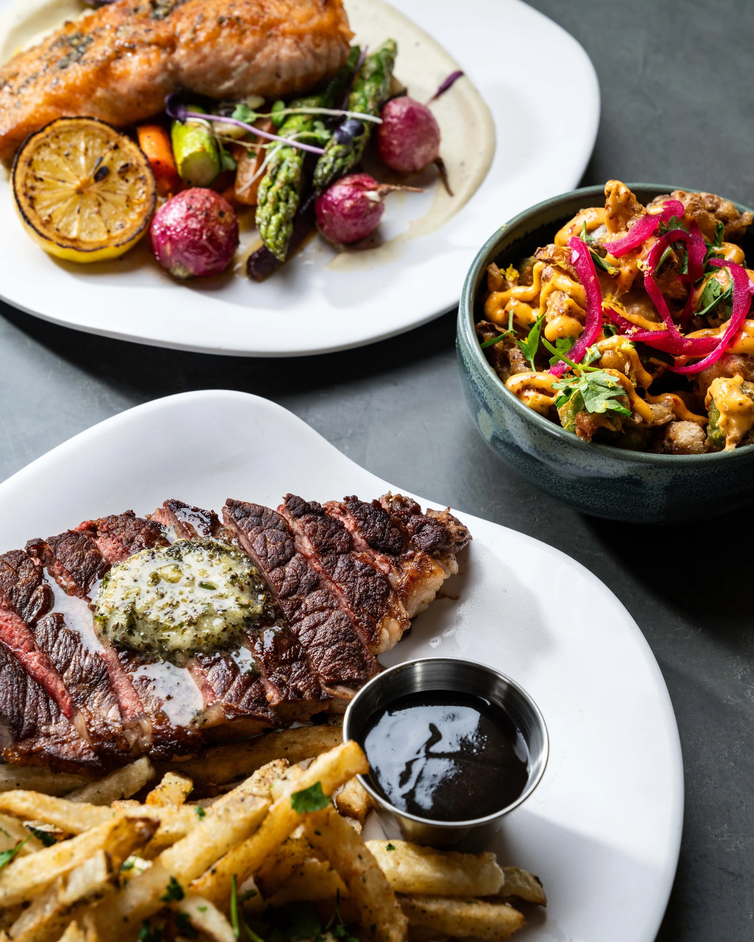 A close-up of a plate with grilled steak topped with herb butter, French fries garnished with parsley, grilled vegetables including lemon, asparagus, and radishes, and a small cup of black sauce. Part of another dish with crispy fried chicken and pink pickled onions is visible in a bowl nearby.