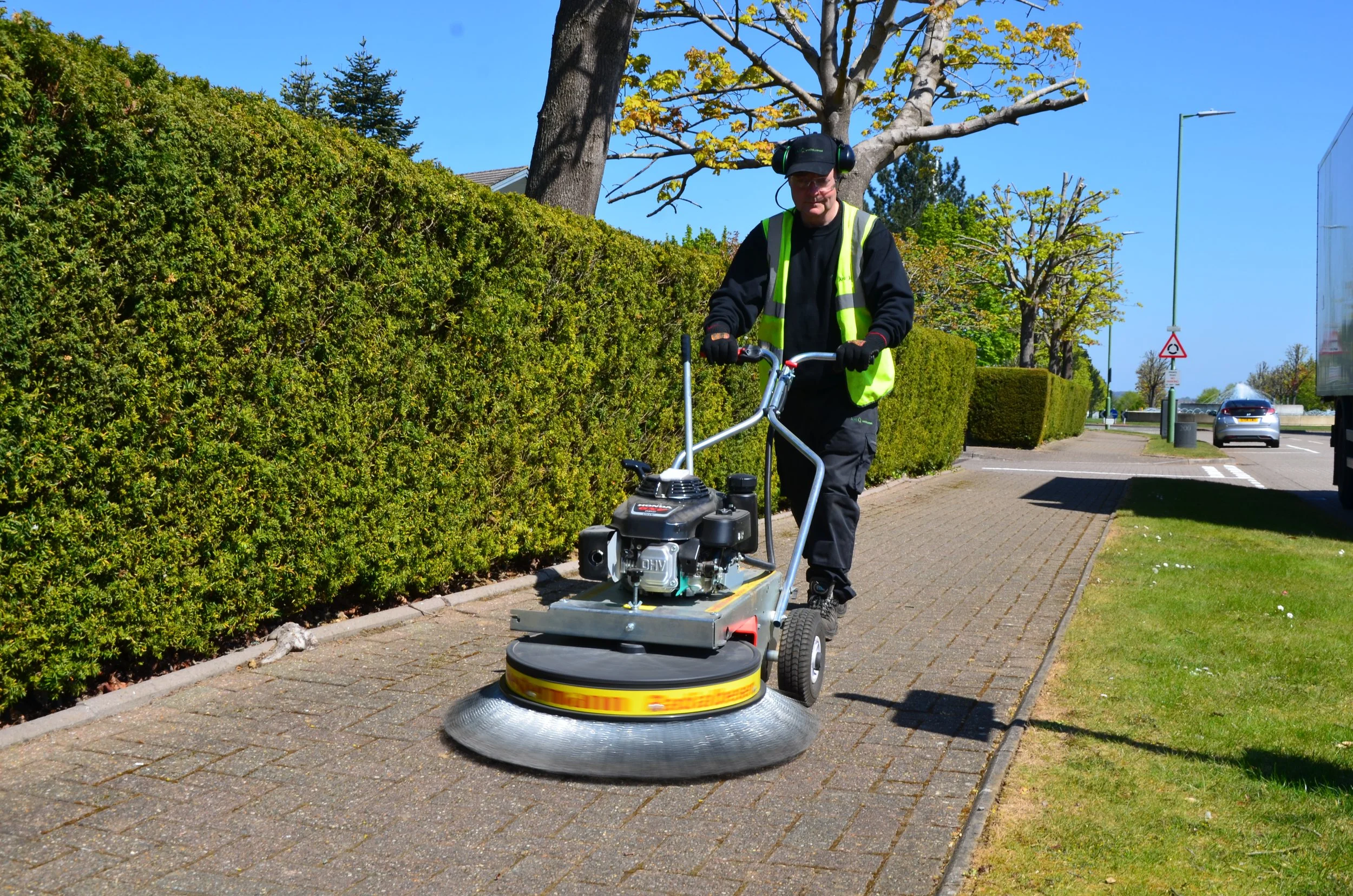 A man in a black shirt and yellow safety vest is operating a sidewalk power trowel on a brick-paved walkway. He is wearing a cap and ear protection, and is pushing the equipment to smooth the pavement. The background shows green bushes, trees, a street sign, parked cars, and a blue sky.