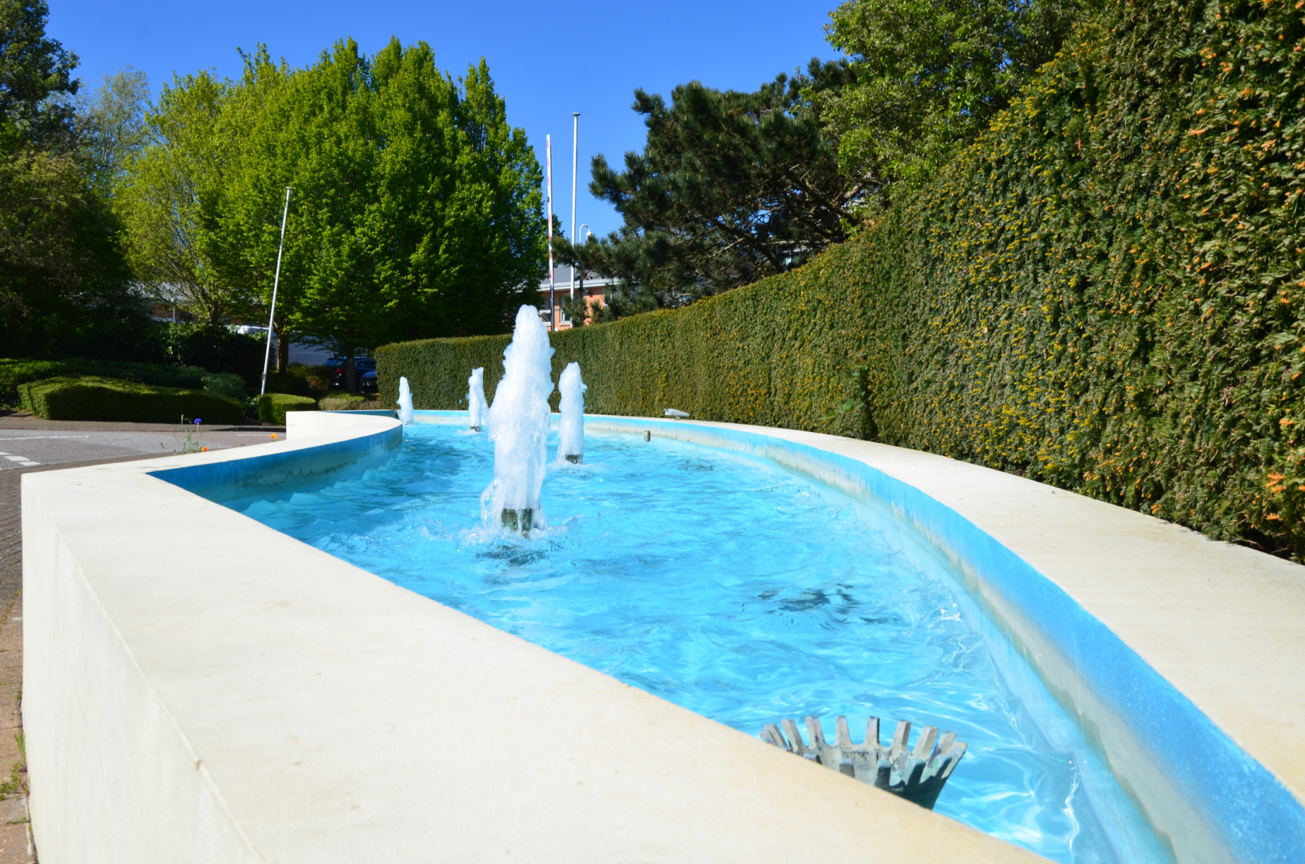 A rectangular water fountain with multiple water jets at Capability Green, Luton surrounded by green trees and a tall, neatly trimmed hedge, under a clear blue sky.