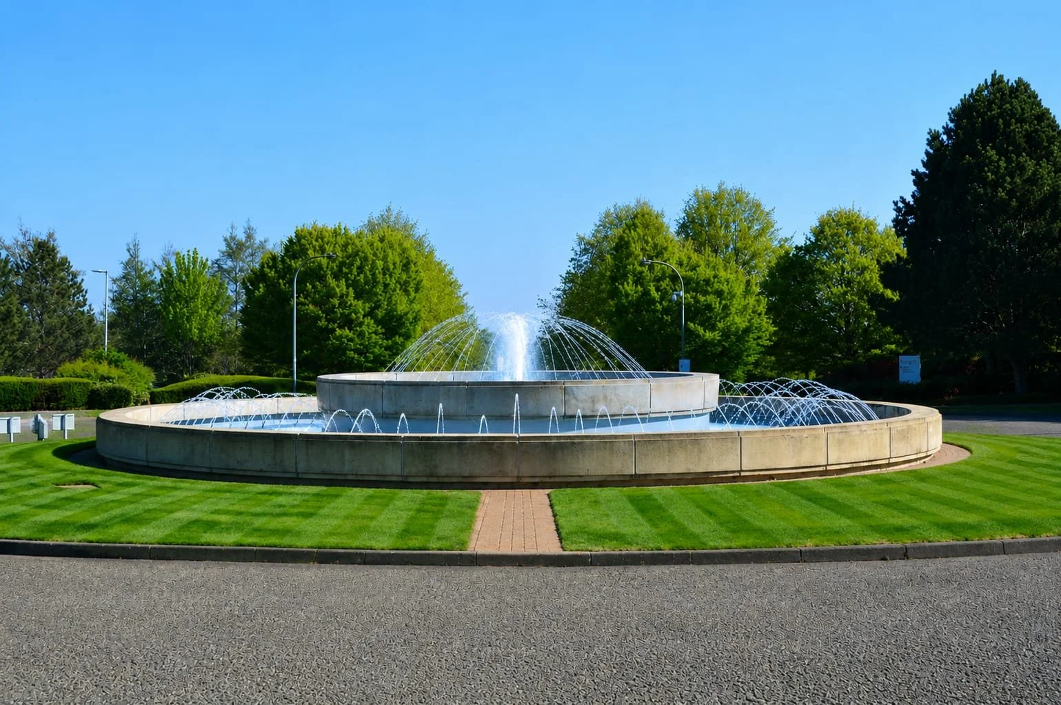Fountain at Capability Green. A large circular stone fountain with water spouting in the center and smaller streams around the edges, surrounded by well-maintained green grass and tall trees under a clear blue sky.
