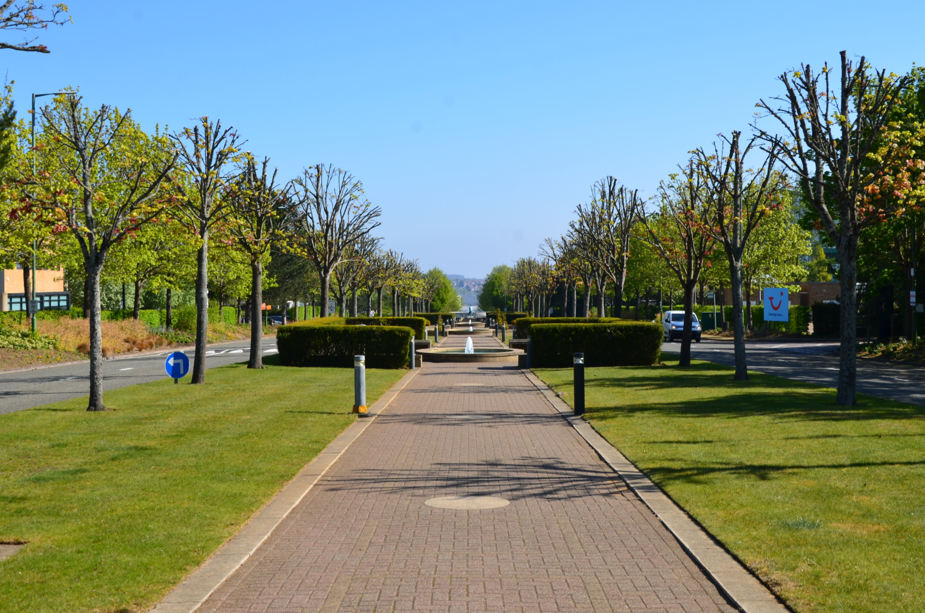A symmetrical park pathway at Capability Green, Luton with trees on both sides and a water fountain in the distance, under a bright blue sky.