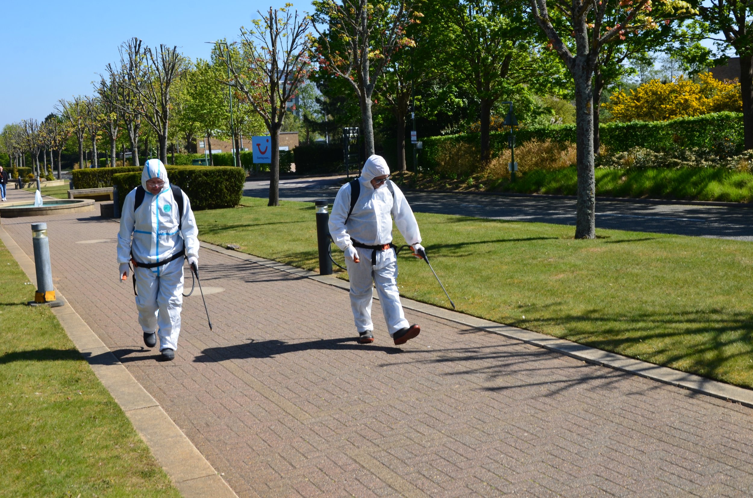 Two AHI Landscaping workers in white protective suits and masks walking outdoors in a park-like area, using handheld equipment for cleaning or spraying, with trees and bushes in the background.