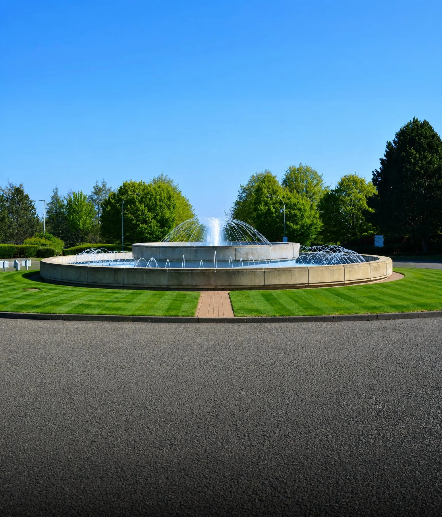 Fountain at Capability Green, Luton. A round fountain with spouting water in a park area, surrounded by green trees and a neatly trimmed lawn, with a part of a paved walkway in the foreground and street lamps nearby.