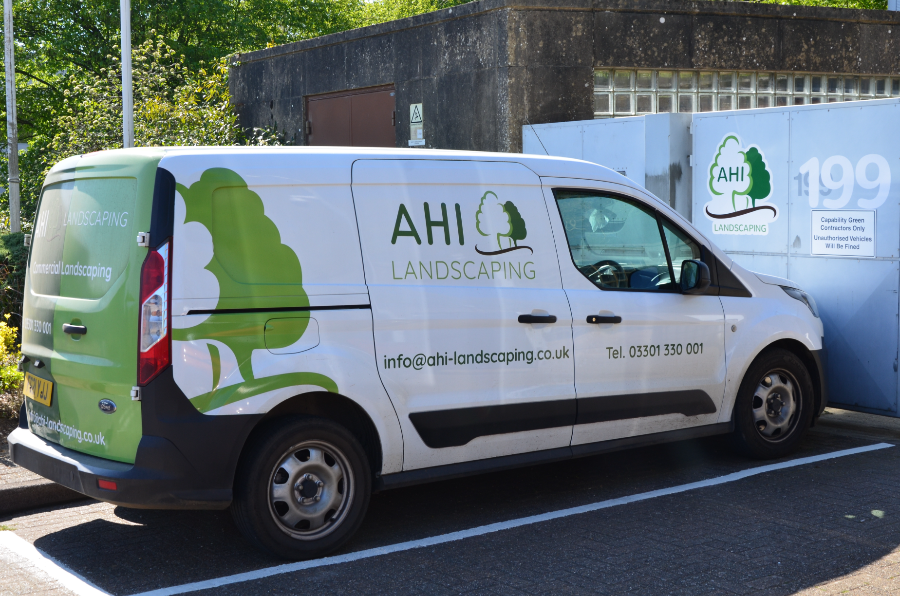 A white van with green and black branding for AHI Landscaping, parked in a parking lot next to a building with glass blocks. The van has a large tree logo and contact information including an email and phone number.