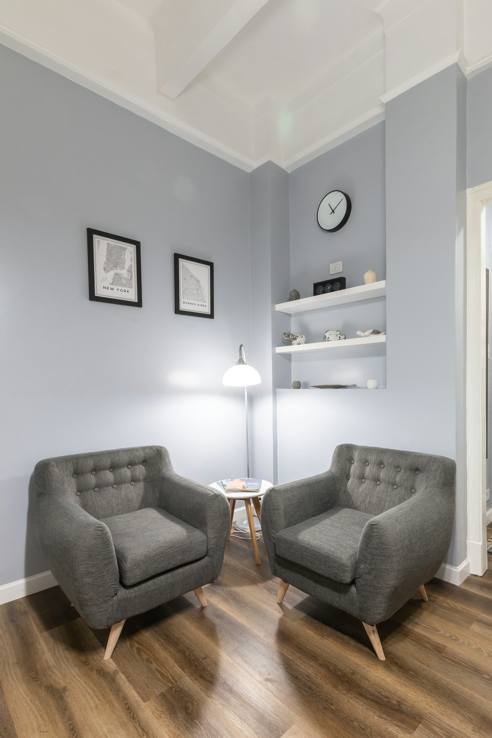 A cozy living room corner with two gray upholstered armchairs facing each other around a small round wooden table with magazines on it. The room has a light blue wall with two framed maps of New York and Buenos Aires. There is a white floating shelf with decorative objects and a black round wall clock. A floor lamp with a white shade provides lighting. The floor is wood with a natural finish.