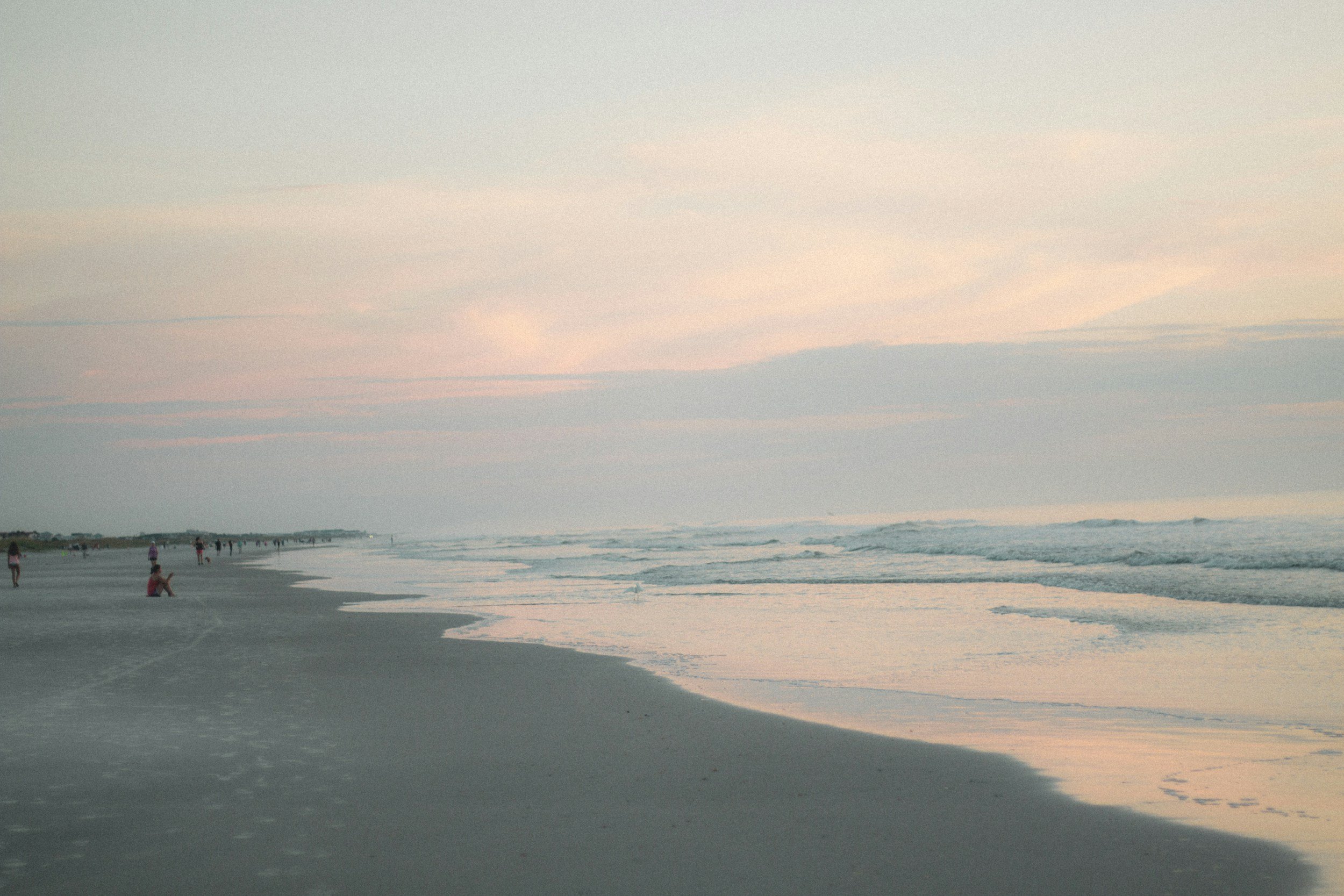 A serene beach scene at dusk with people strolling and sitting on the sand, calm ocean waves, and a pastel-colored sky with soft clouds.