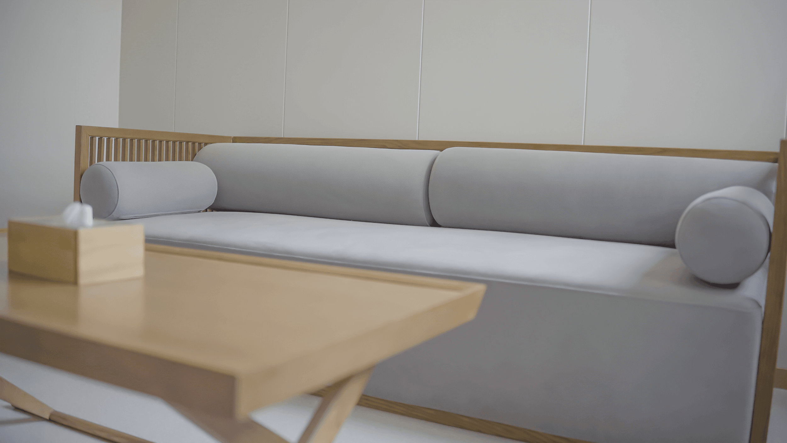 A minimalist waiting area with a wooden-framed sofa with white cushions and cylindrical pillows, and a light wood coffee table with a tissue box on top.