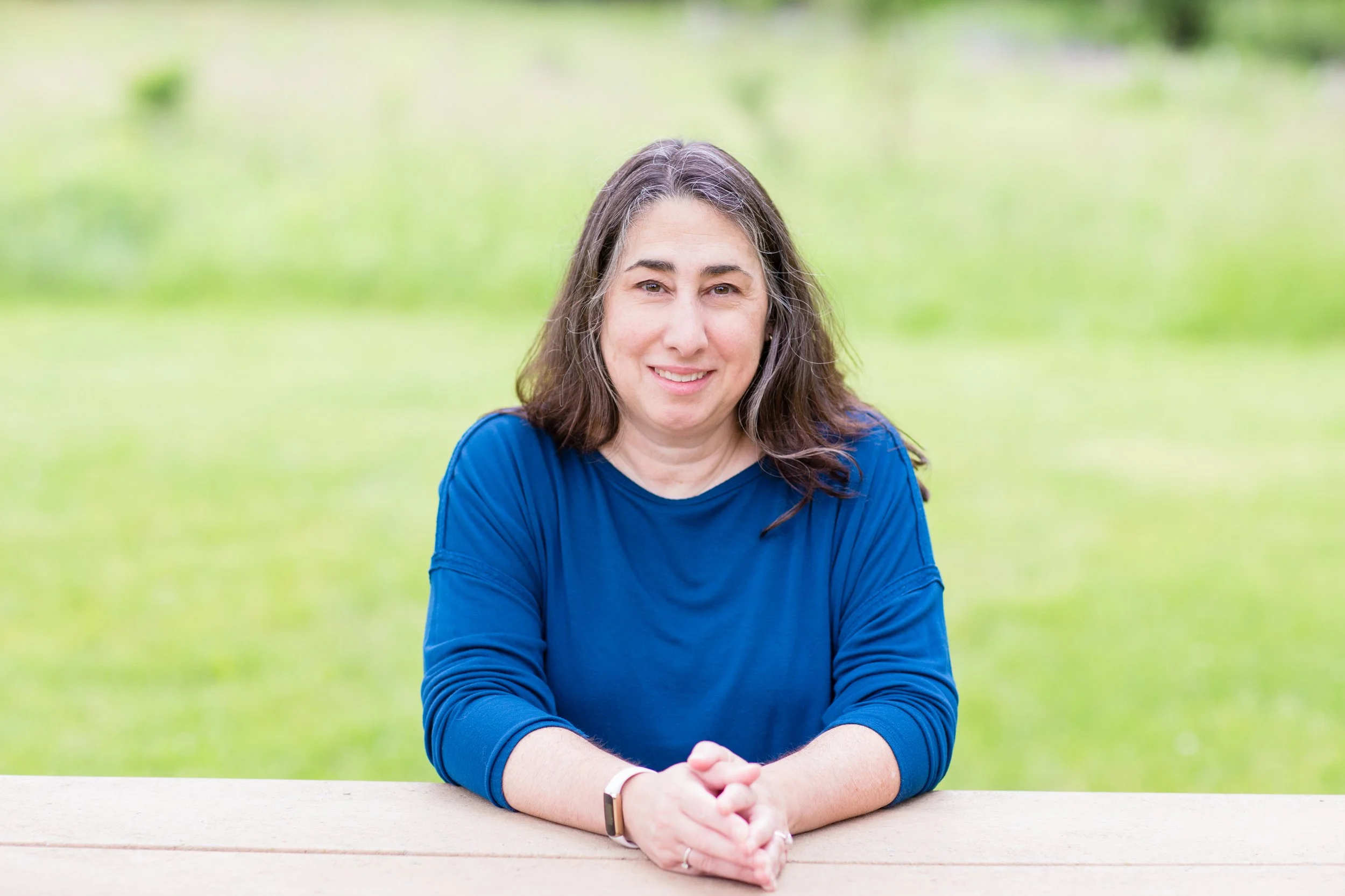 A woman with shoulder-length gray hair wearing a blue long-sleeve shirt, sitting outdoors at a wooden table with her hands clasped, smiling at the camera, with a blurred green grassy background.