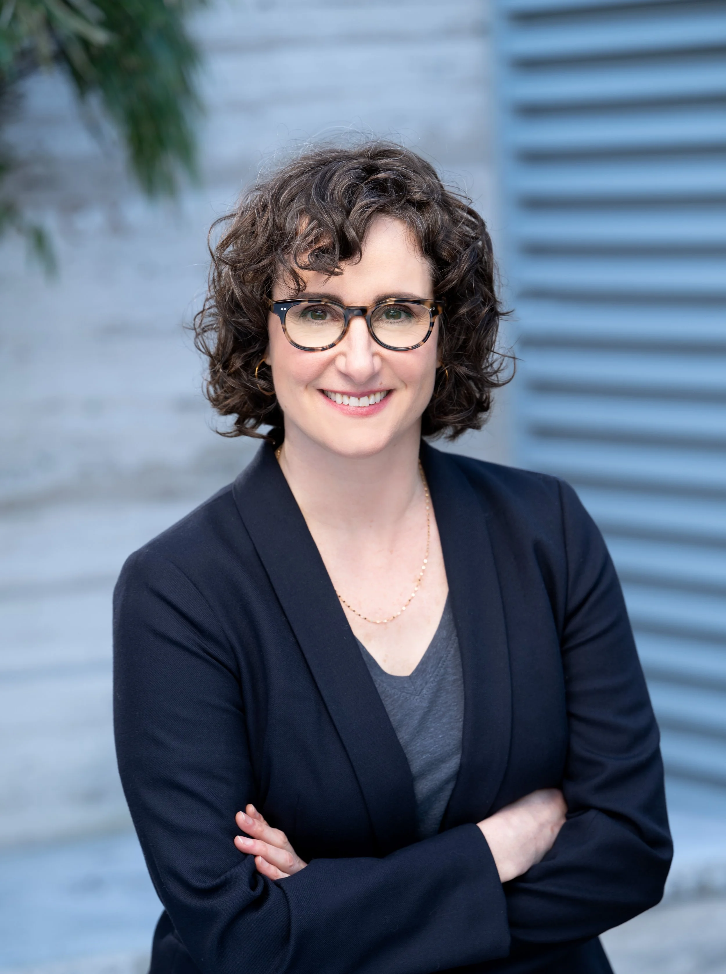 A woman with curly brown hair, wearing glasses, a black blazer, and a gray top, stands outdoors with her arms crossed, smiling at the camera.