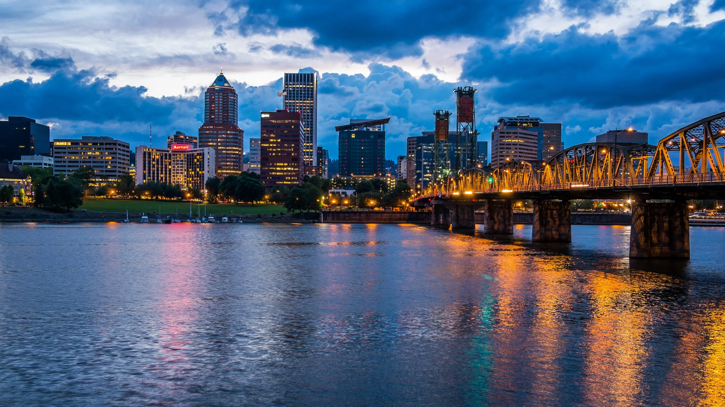 City skyline at dusk with illuminated skyscrapers, a bridge over water, and a cloudy sky.
