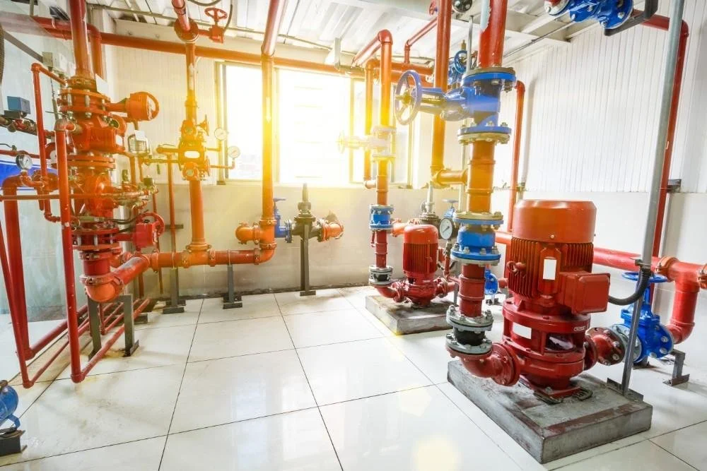 Industrial pump system with red and blue pipes and valves in a facility with sunlight coming through the window.