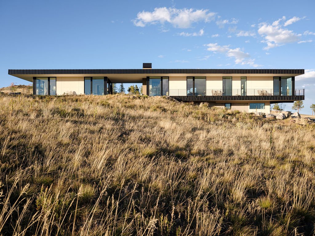 Modern house with large glass windows and a balcony, situated on a grassy hillside under a partly cloudy sky.