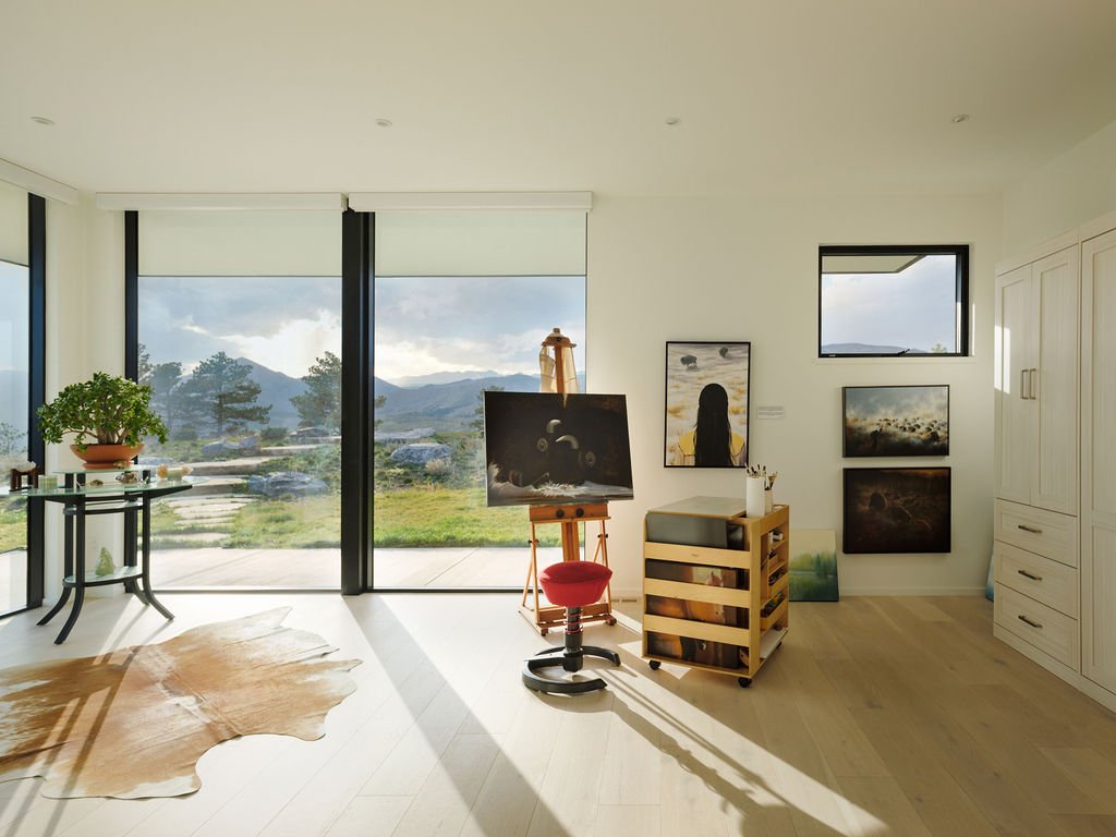 Bright art studio with large glass sliding doors showcasing a mountain landscape, an easel with a painting, framed artwork on the wall, a wooden storage cart, and a potted plant on a small table.