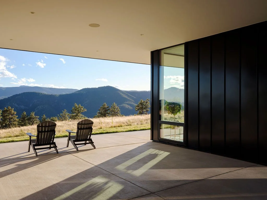 A covered outdoor patio with two wooden chairs overlooking a scenic mountain landscape, with trees and blue skies.