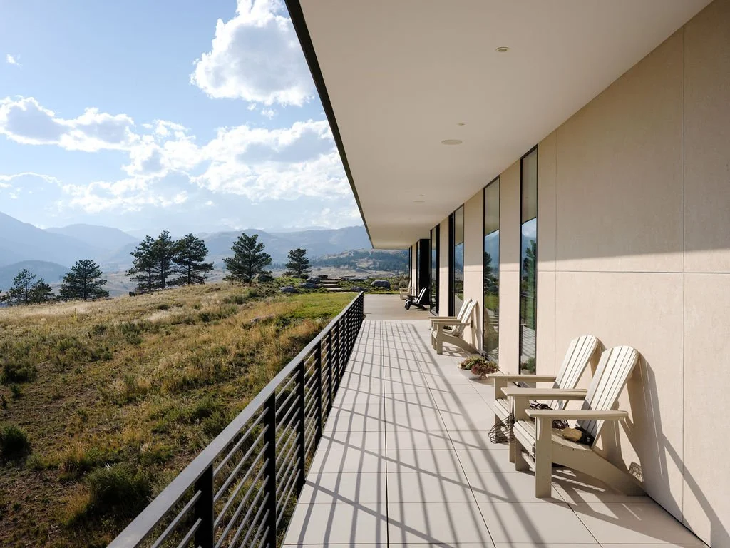 A spacious balcony with chairs, overlooking a grassy field with trees and mountains in the distance under a partly cloudy sky.