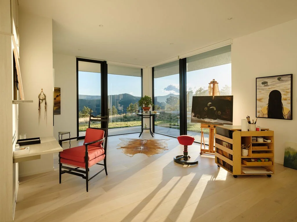 Bright art studio with large floor-to-ceiling windows showcasing a mountain landscape, containing a red lounge chair, an easel with a painting, a rolling art supply cart, and various artworks.