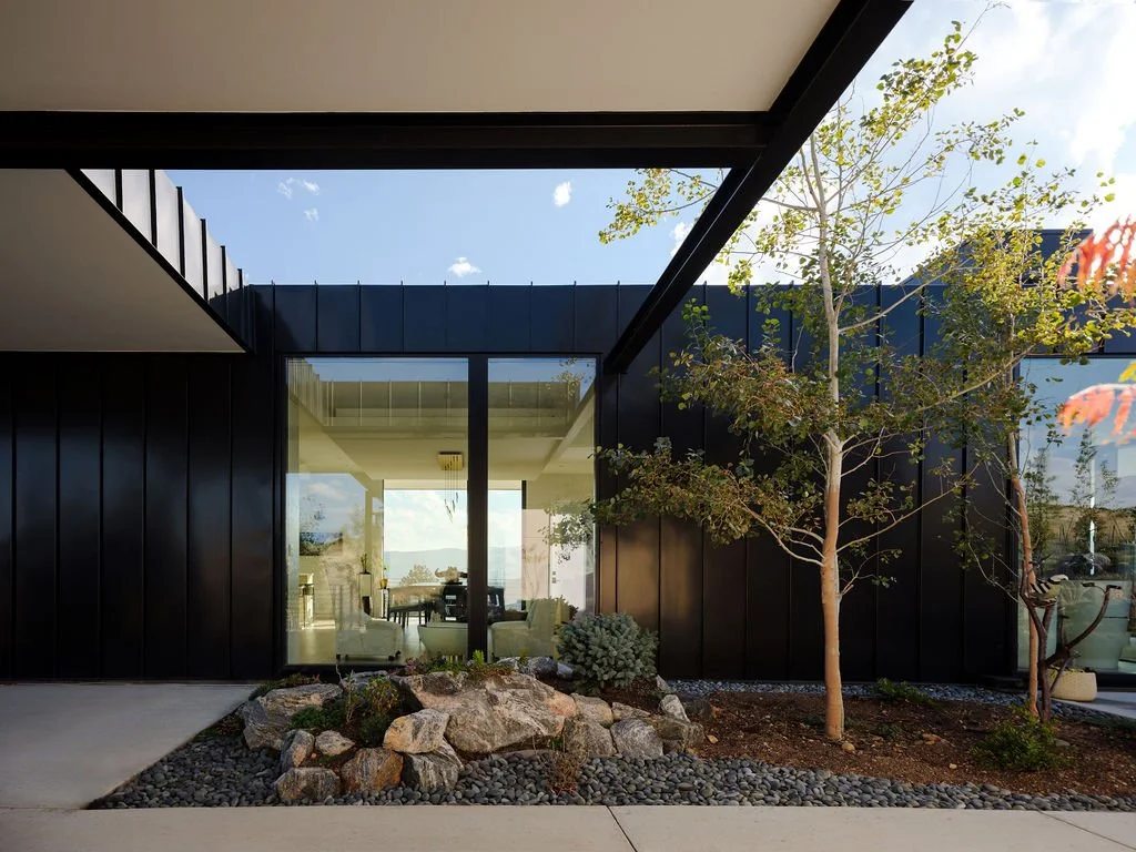 Exterior view of a modern building with black metal siding, large glass windows, and a landscaped front yard with rocks, trees, and shrubs.