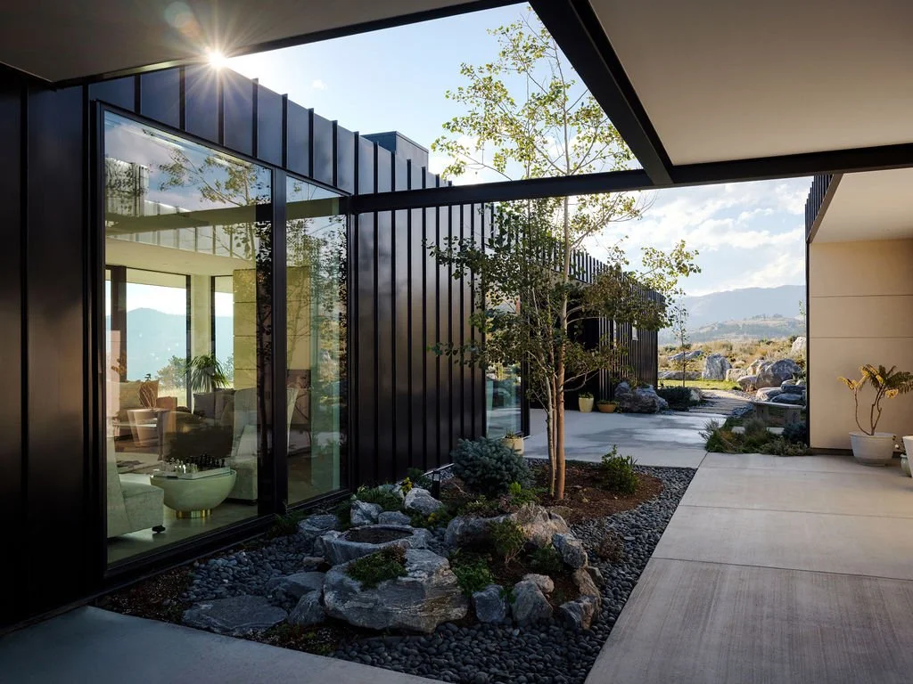 Modern house exterior with large glass windows and a small landscaped area with rocks and trees, overlooking a scenic landscape with hills and rocks, under a clear sky.