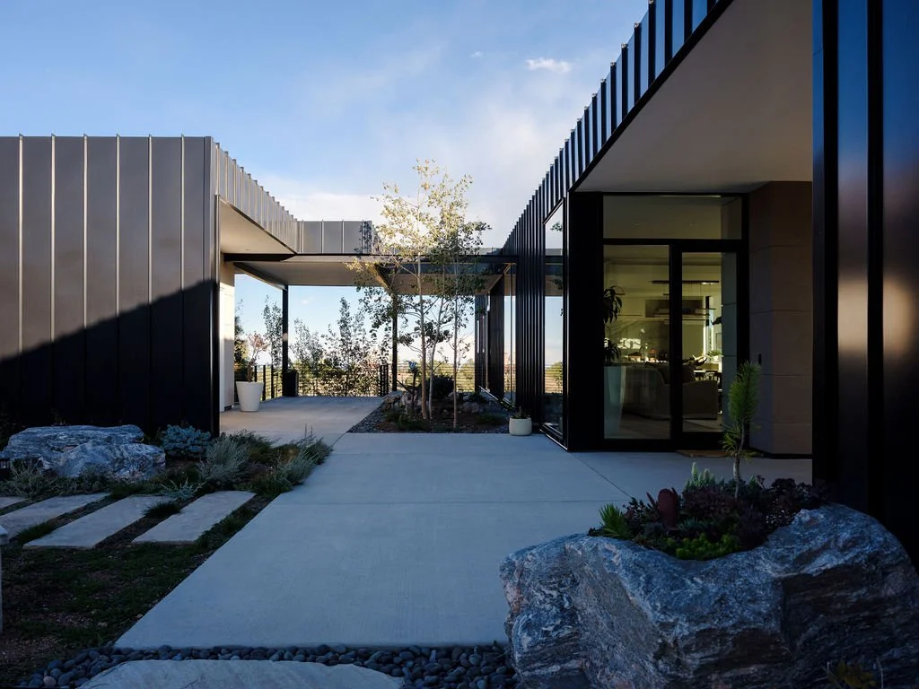 Modern building exterior with black metal siding, large glass doors, and a concrete walkway surrounded by rocks, plants, and trees.