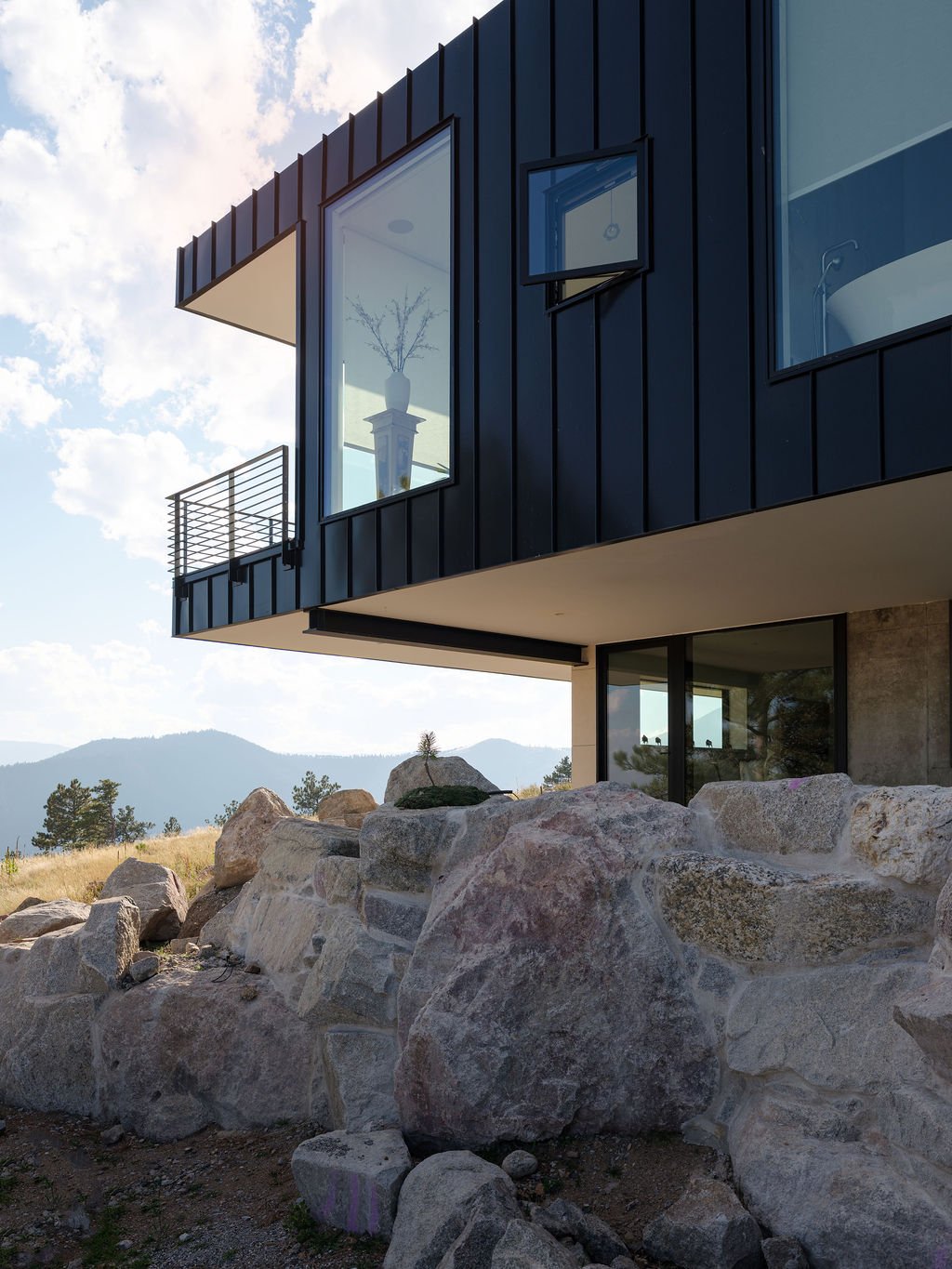 Modern black house with large windows and a balcony, set on rocky terrain with mountain views in the background.