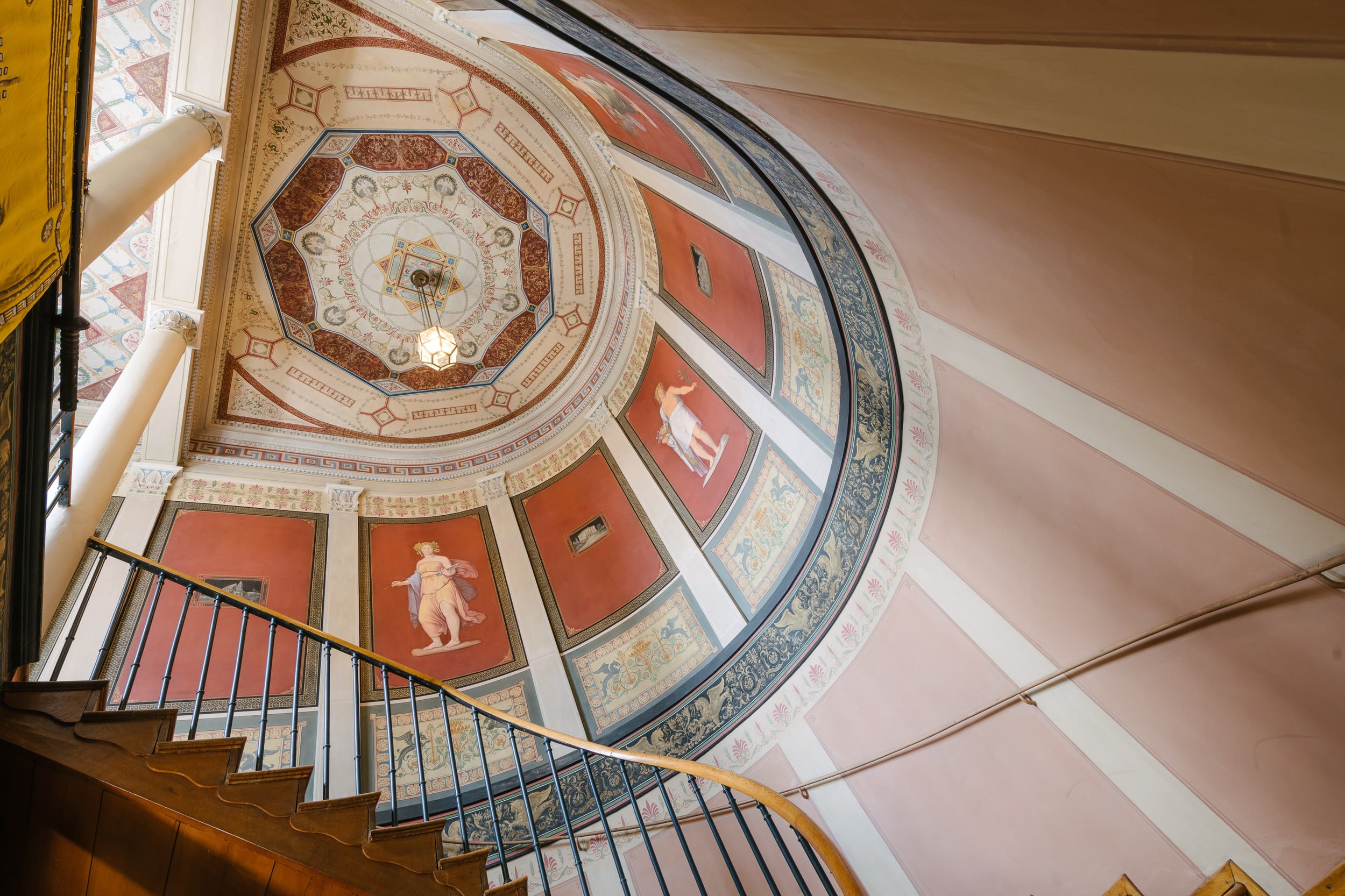 Die Decke und Wände dieses Stiegenhauses sind mit Fresken in Rot-, Beige- und Blautönen bemalt. Die Wände darunter sind mit rosa Farbe gestaltet.