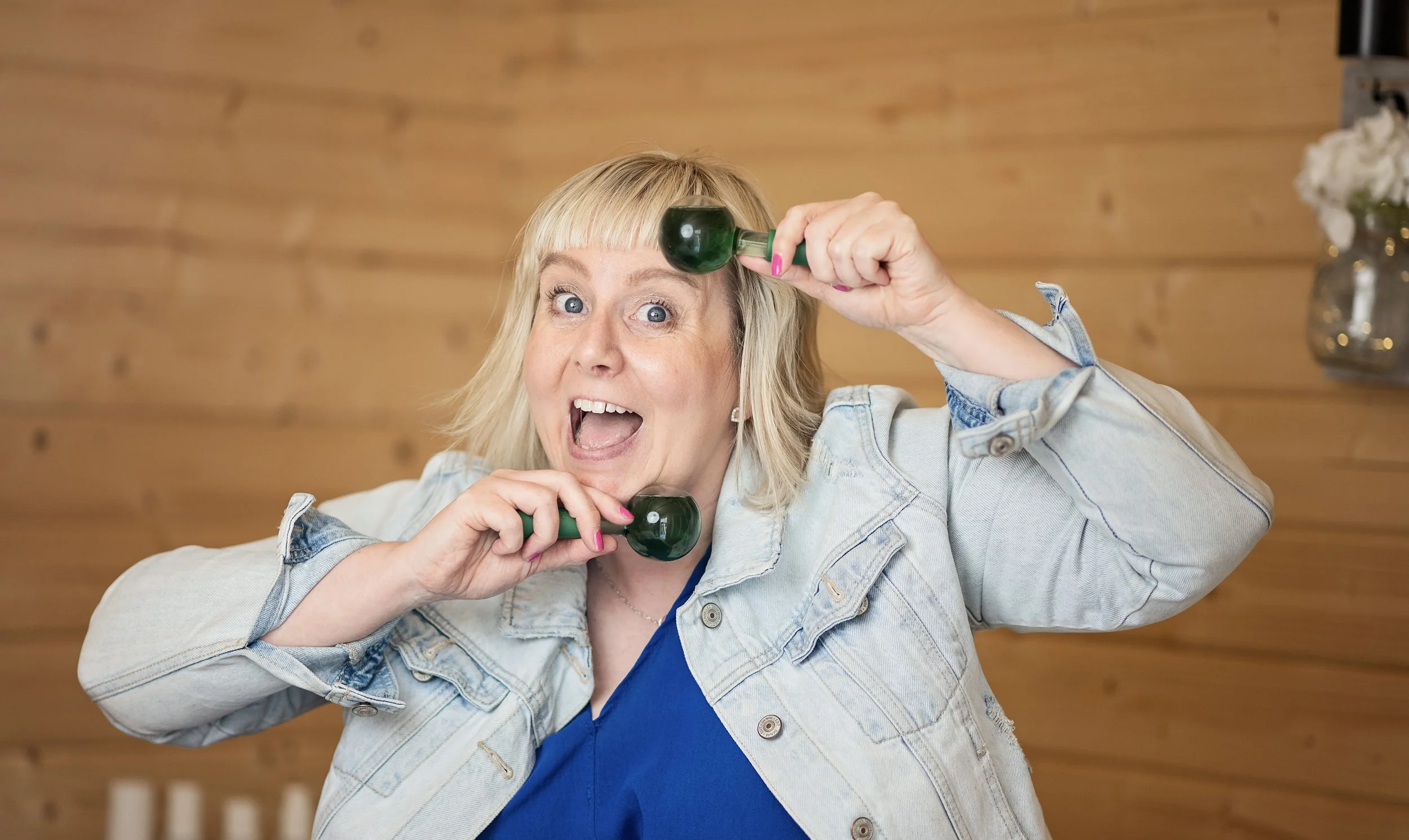A middle-aged woman with blonde hair and blue eyes holding green maracas near her head with an excited expression in a room with a wooden wall and a decorative vase in the background.