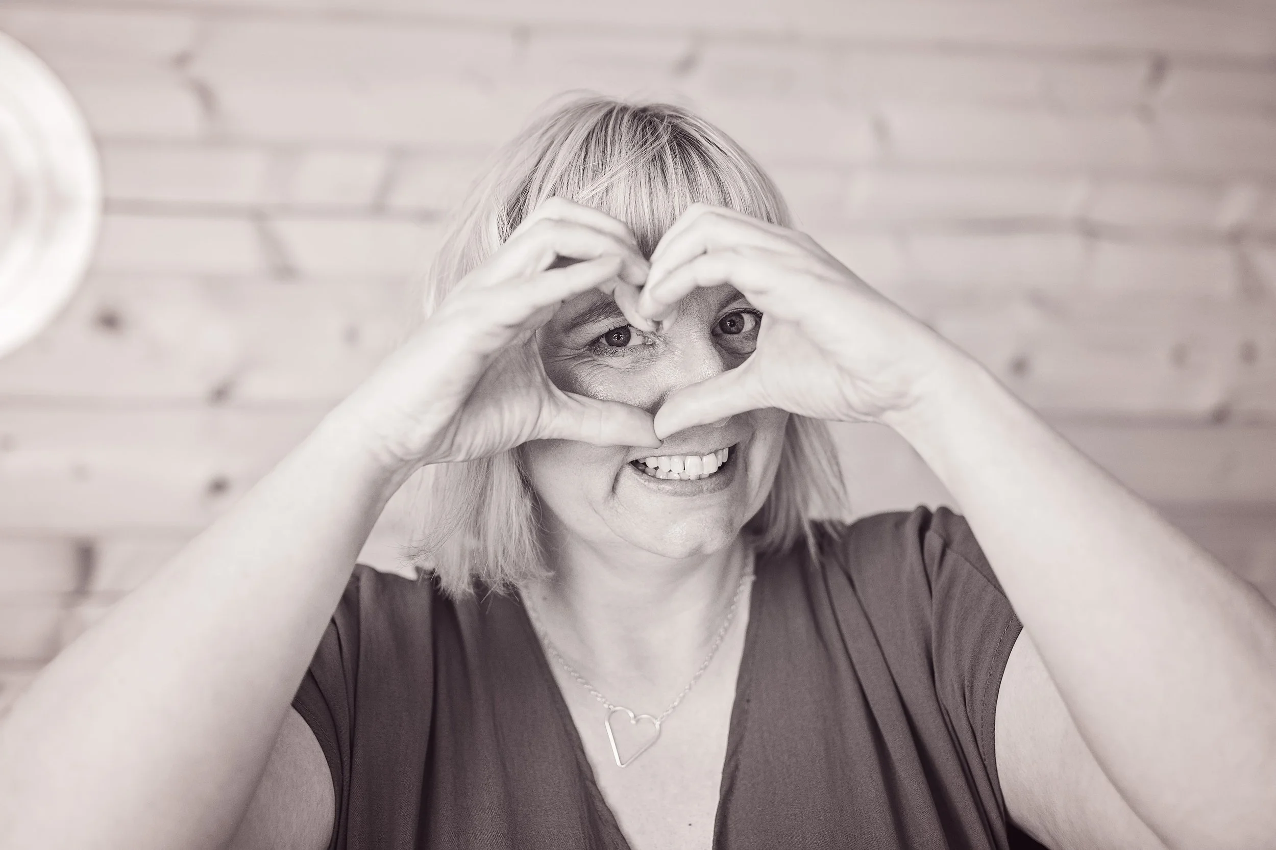 A smiling older woman with blonde hair making a heart shape with her hands in front of her face.