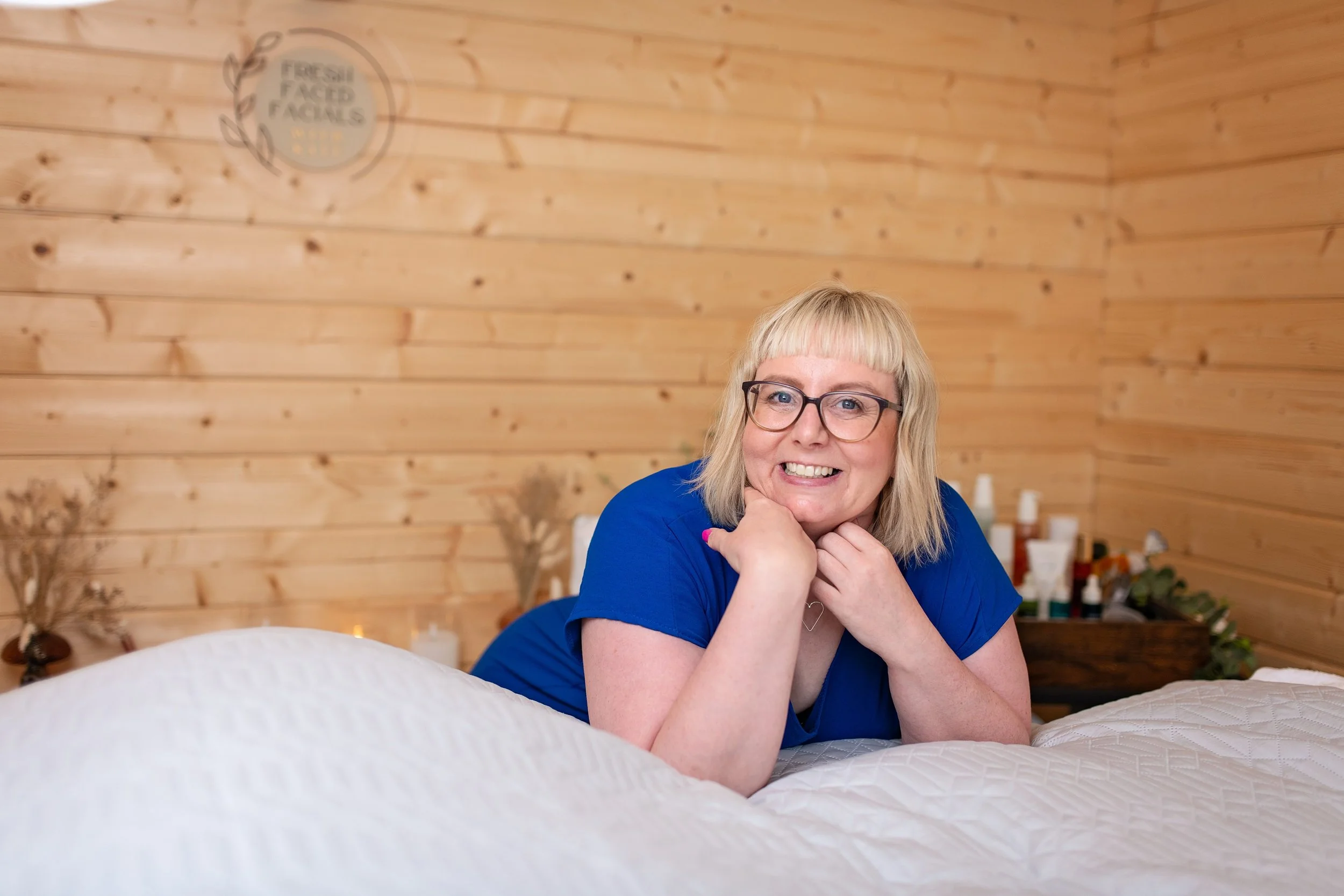 A smiling woman with blonde hair, glasses, and a blue shirt lying on a bed with a wooden wall background, resting her chin on her hands.