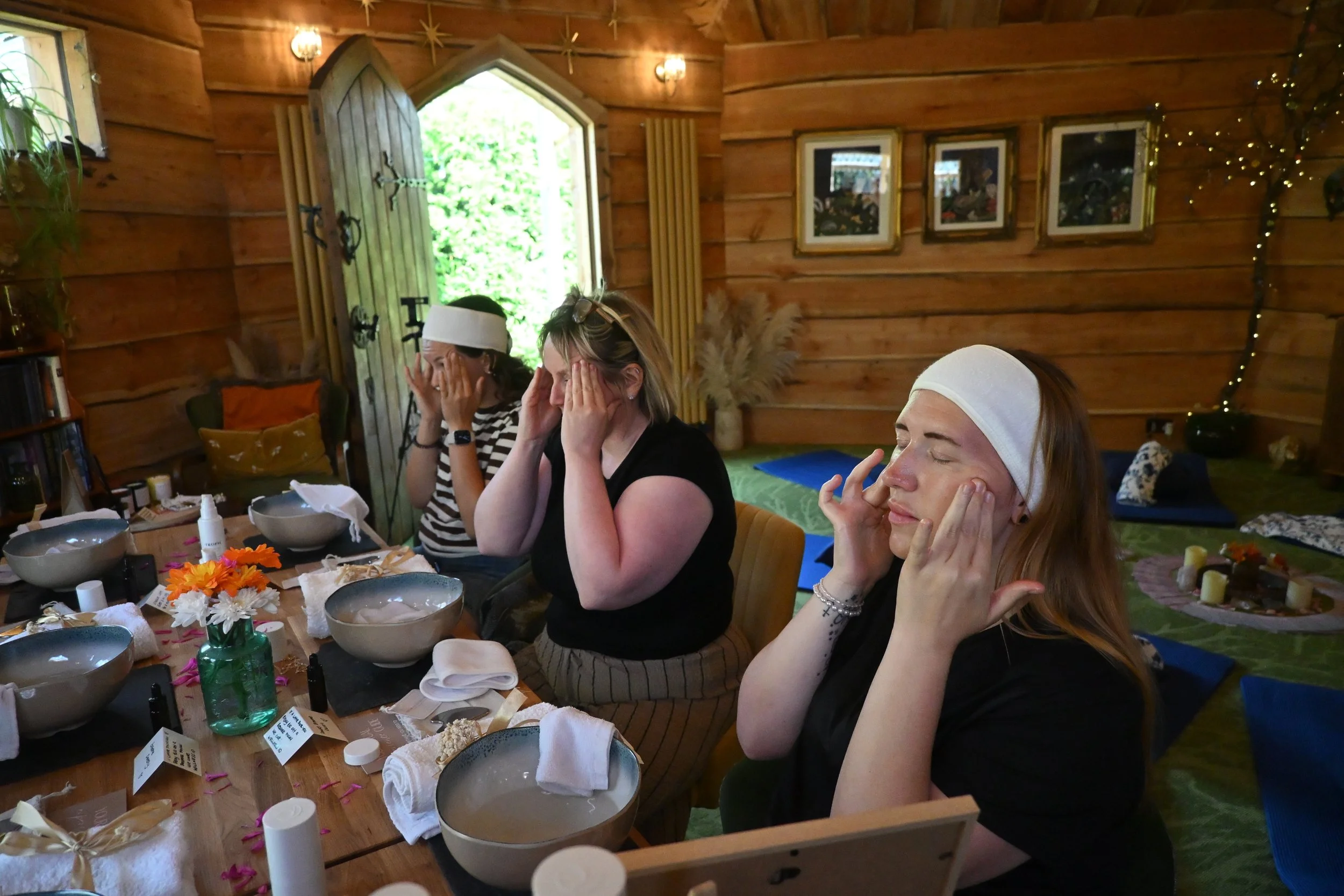 Three women sitting at a wooden table with bowls, candles, and flowers, inside a cozy wooden cabin with framed pictures on the wall, greenery outside, and string lights.