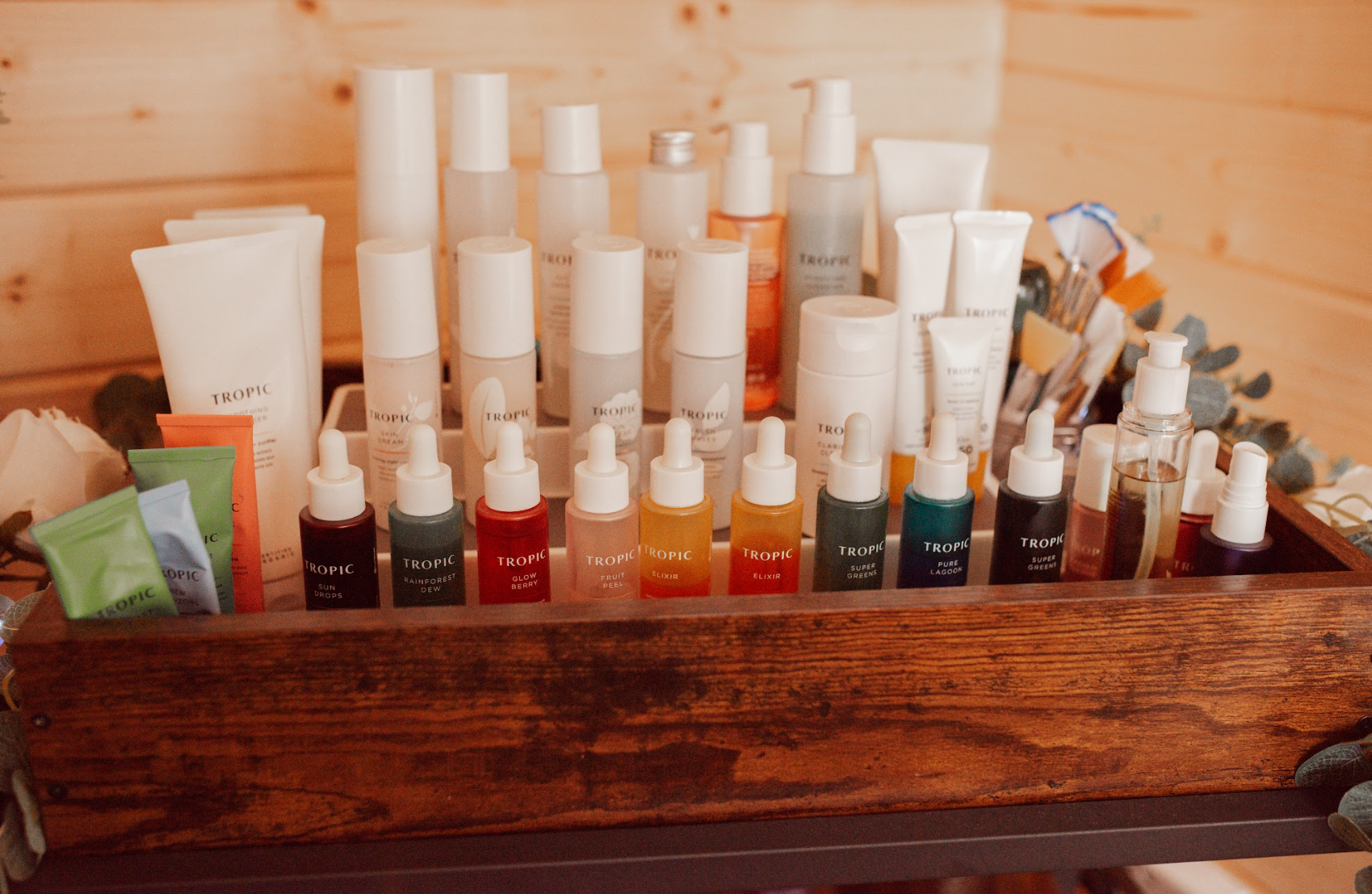 A wooden tray holding a variety of skincare products including bottles, tubes, and droppers, with some labeled Tropic and others with different brands and colors, on a wooden surface against a wood-paneled wall.