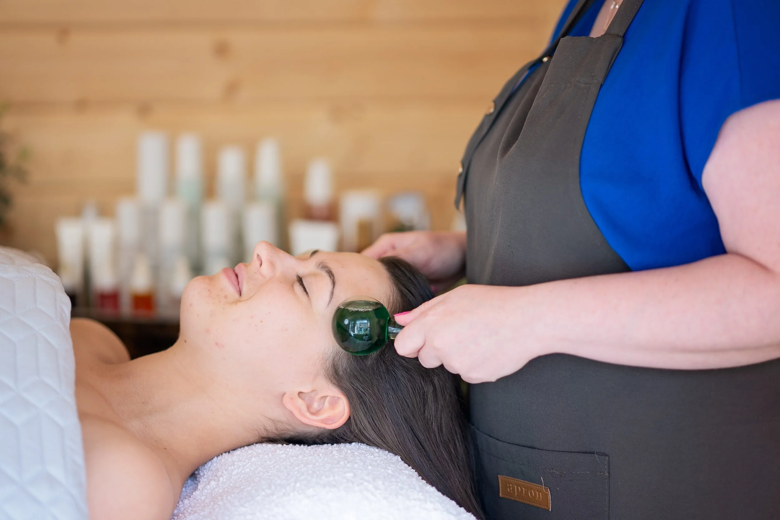 A woman lying down with eyes closed receiving a facial treatment with a glass suction device on her cheek in a spa or wellness center.