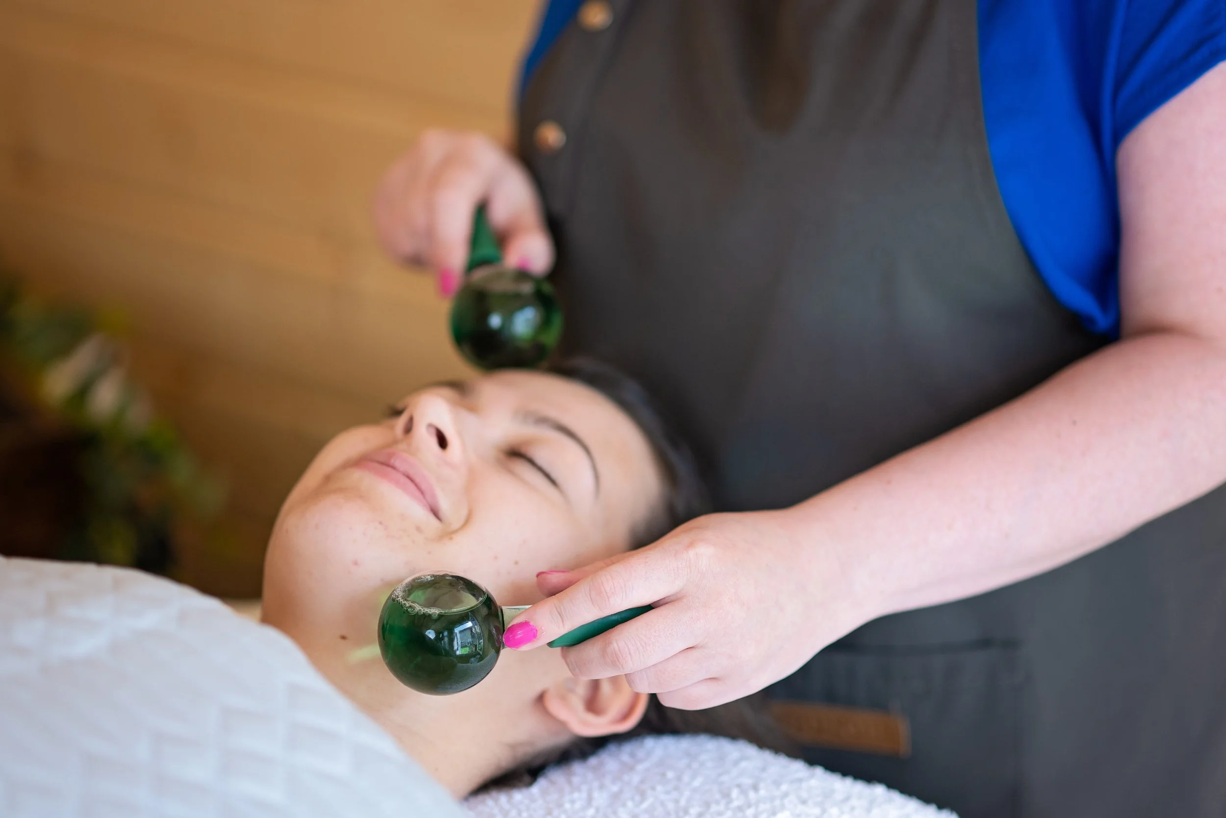 A woman receiving a facial massage with jade rollers while lying down with her eyes closed.