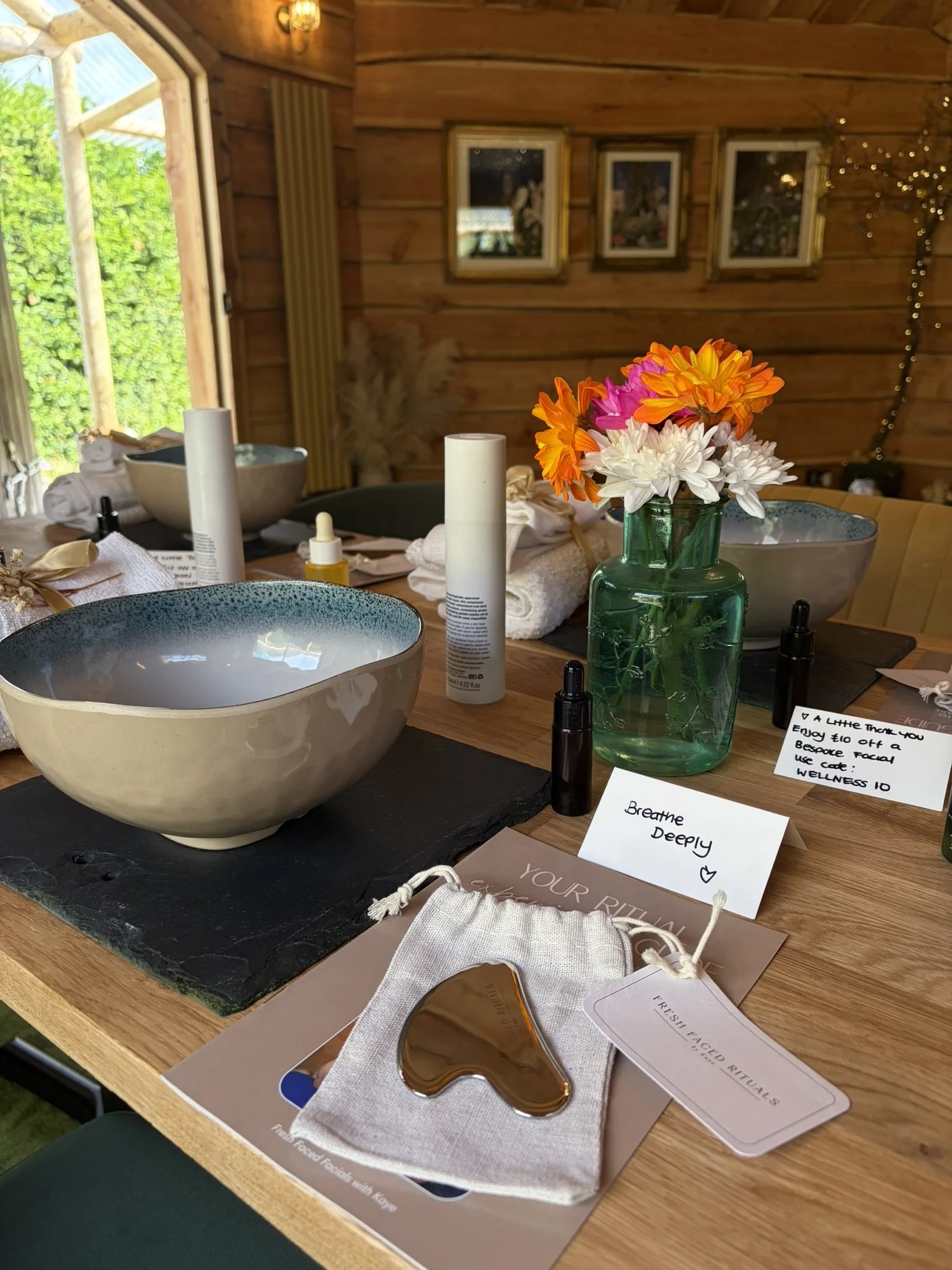 A wooden table with bowls, bottles, a box, a mirror, and a colorful bouquet of flowers in a green glass vase. There are various beauty and skincare products, notes, and a gold-colored facial roller. The background features a wooden wall with framed artwork and a window showing greenery outside.