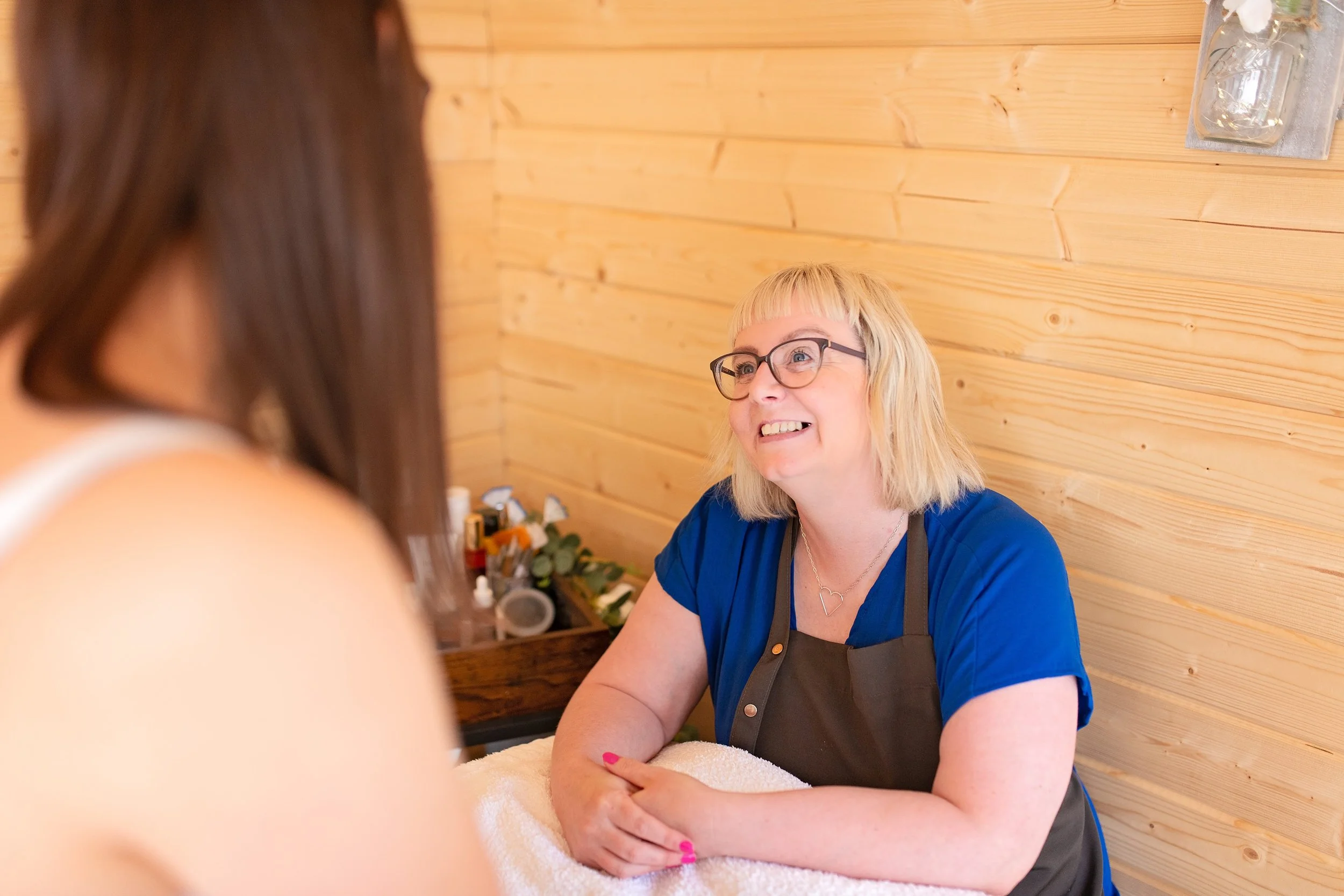 A woman with blonde hair, glasses, and a black apron smiling at a person, in a wood-paneled room. She is giving a massage, resting her arms on a towel-covered surface.